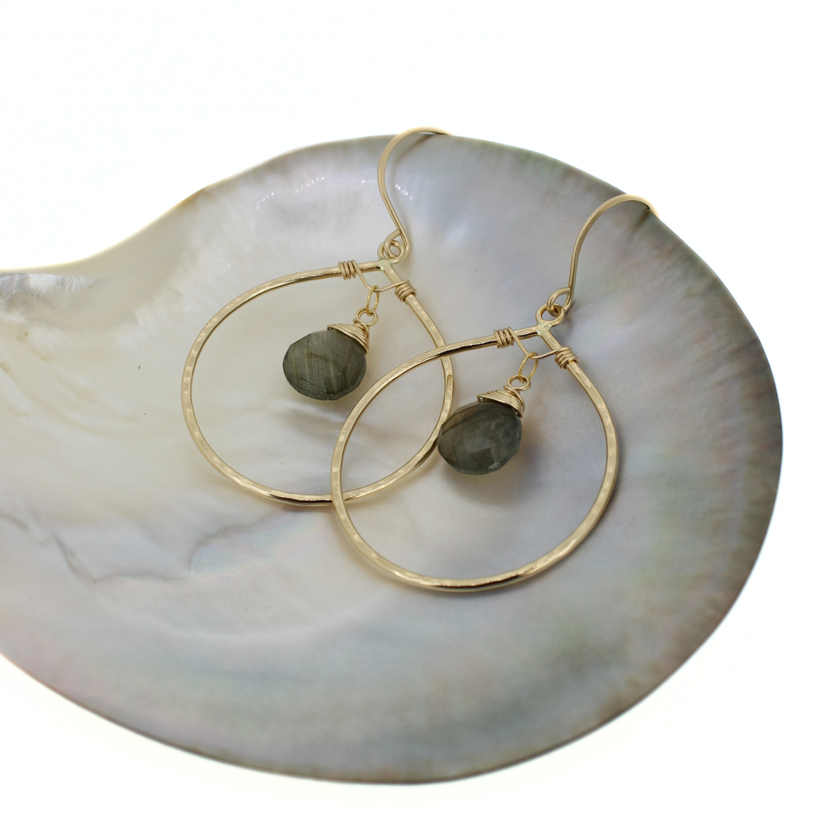 A pair of gold hoop earrings with small dark gray labradorite stones, displayed on a white iridescent shell dish against a white background.