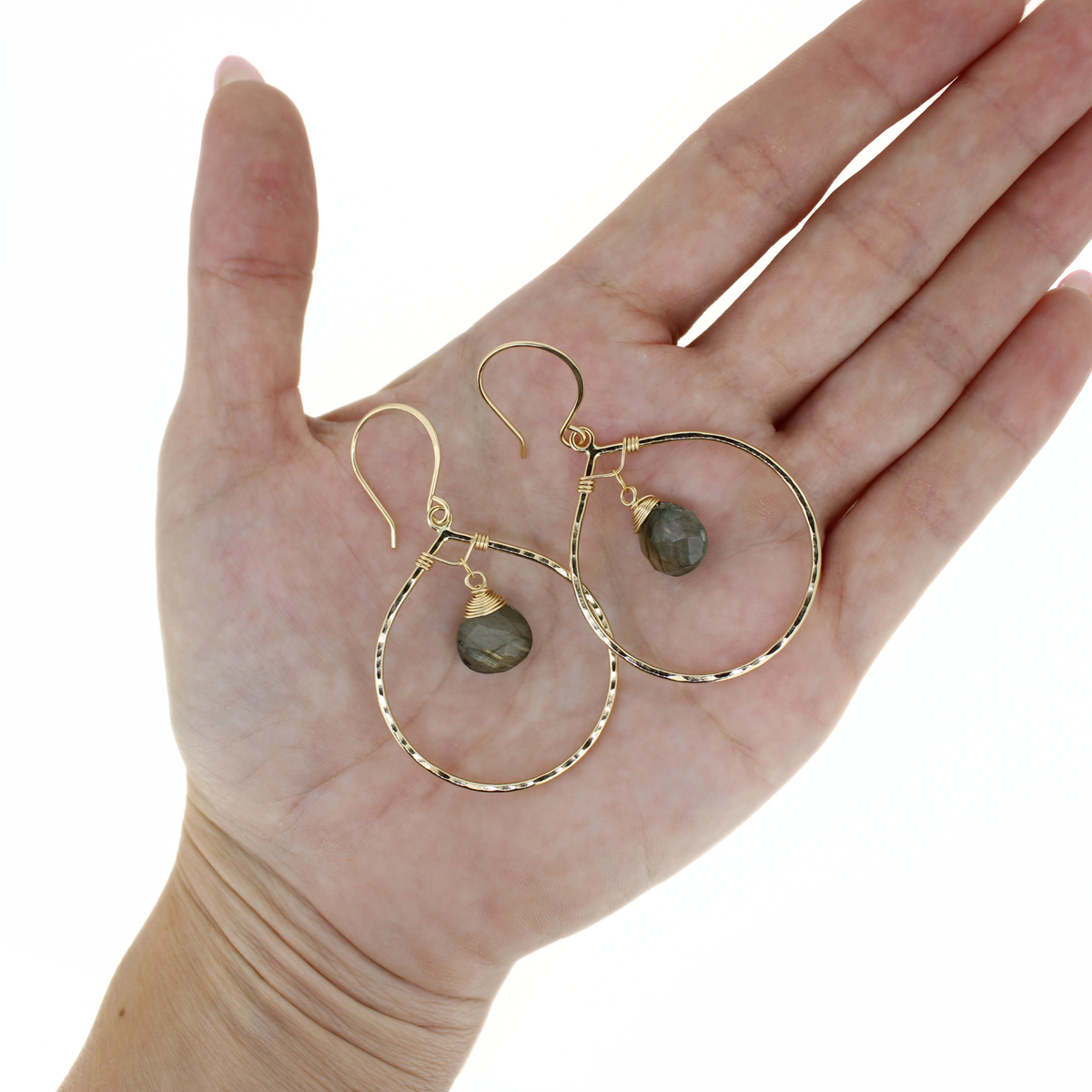 A pair of gold hoop earrings with small faceted gray-green labradorite stone drops, displayed on an open hand against a white background.