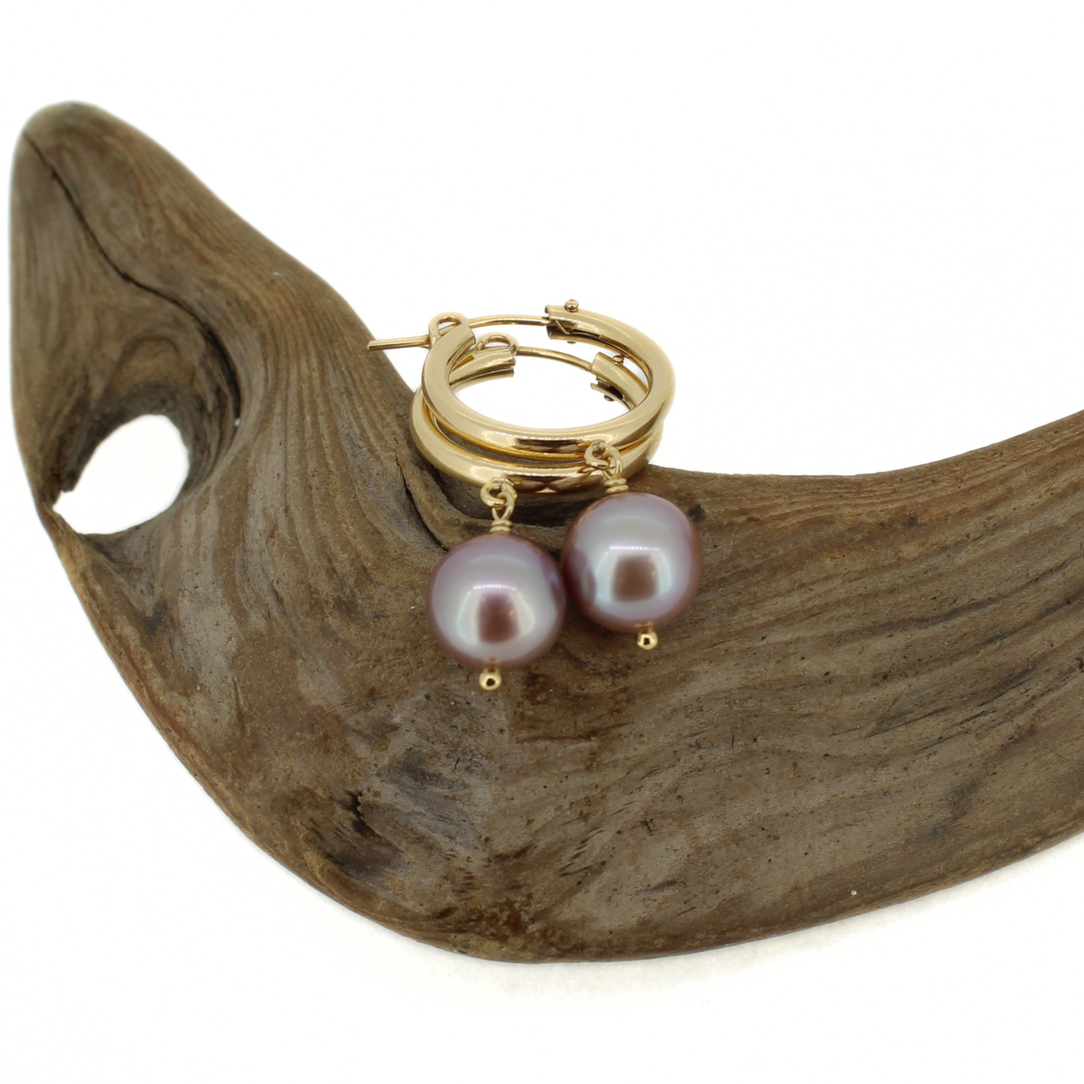 A pair of gold hoop earrings with dangling round pearls in a muted lavender-purple hue, displayed on a curved piece of natural driftwood against a white background.