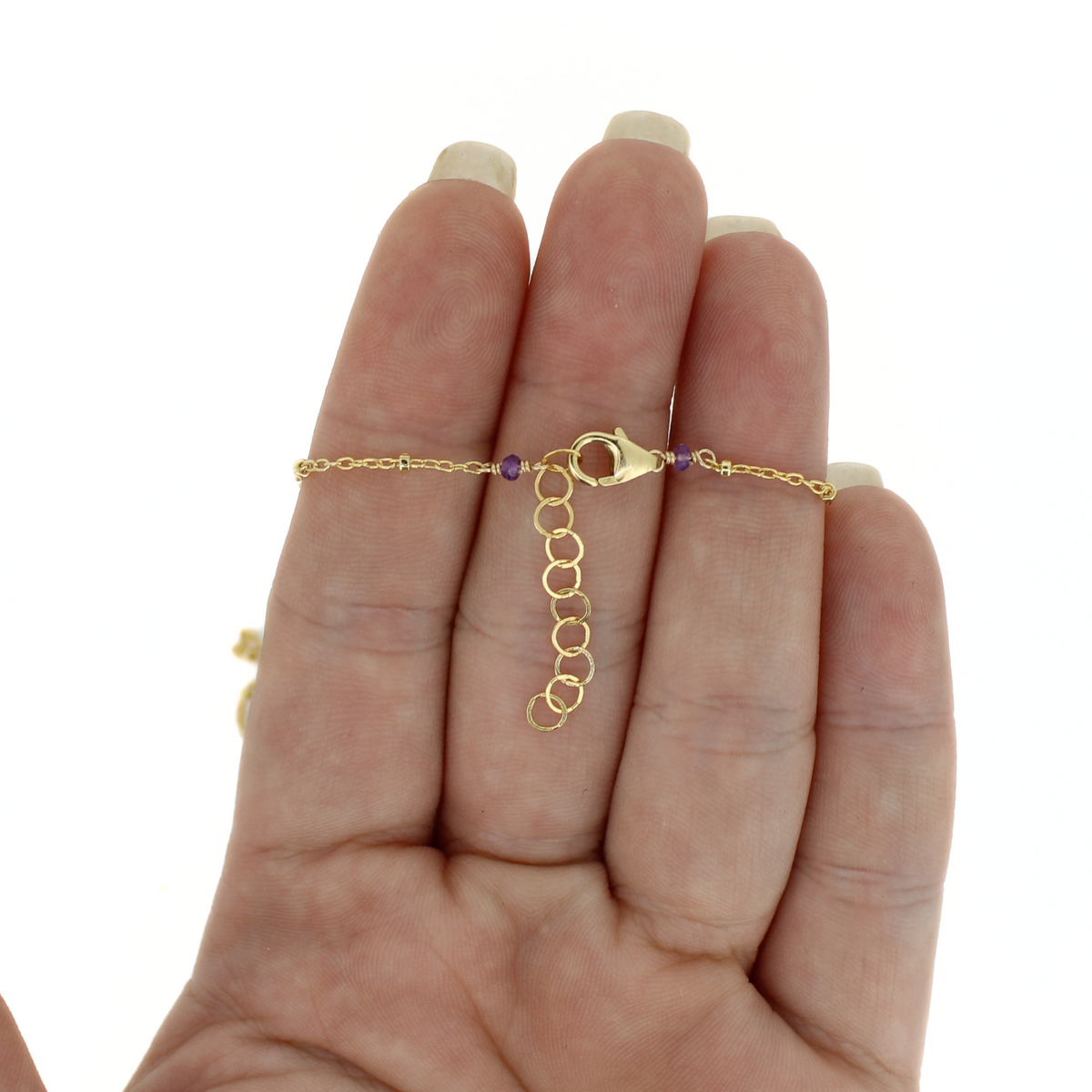A delicate gold chain bracelet with a lobster clasp and purple accent beads, held on a person's fingers against a white background.