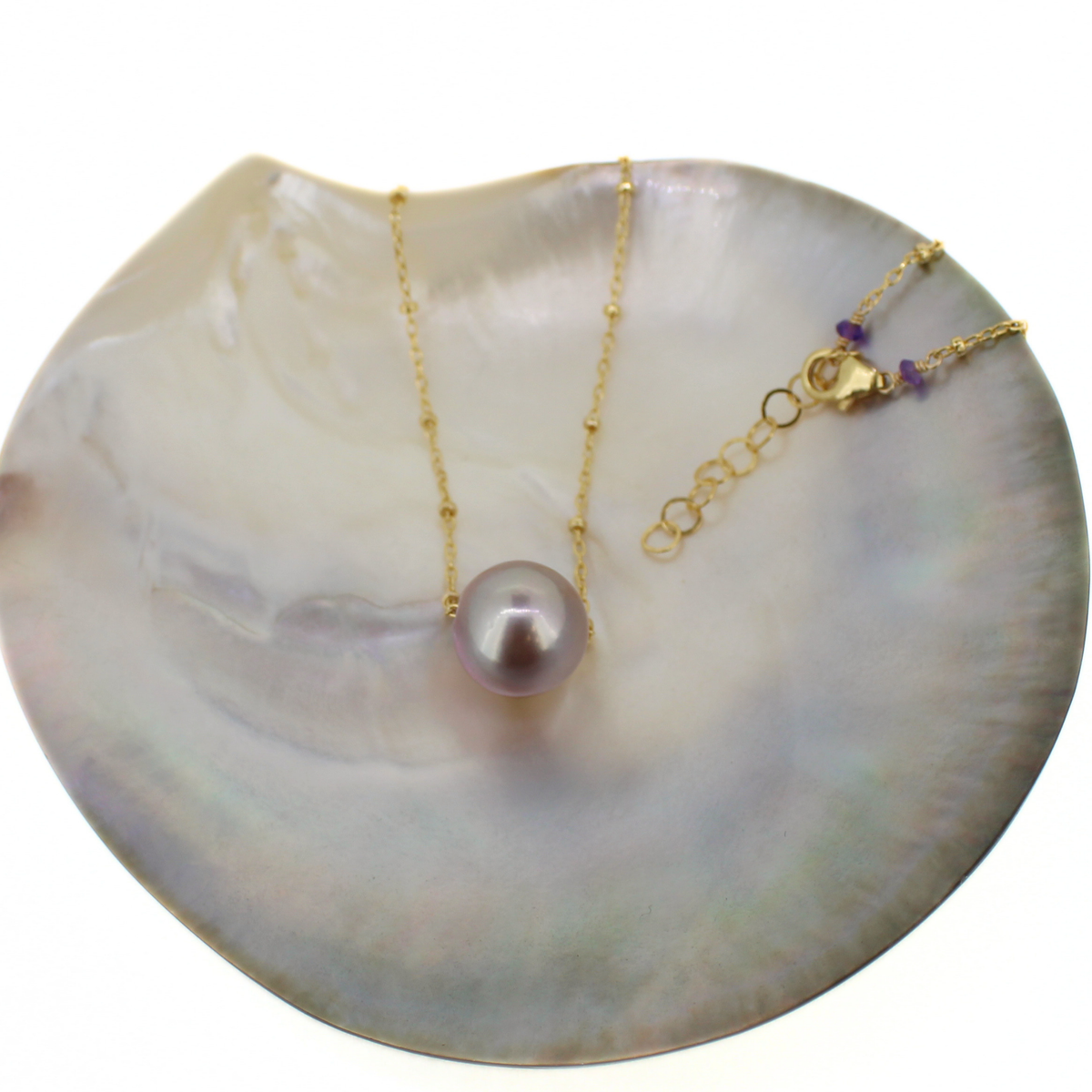 A gold chain necklace with a single round pearl pendant, displayed on a large iridescent shell against a white background.