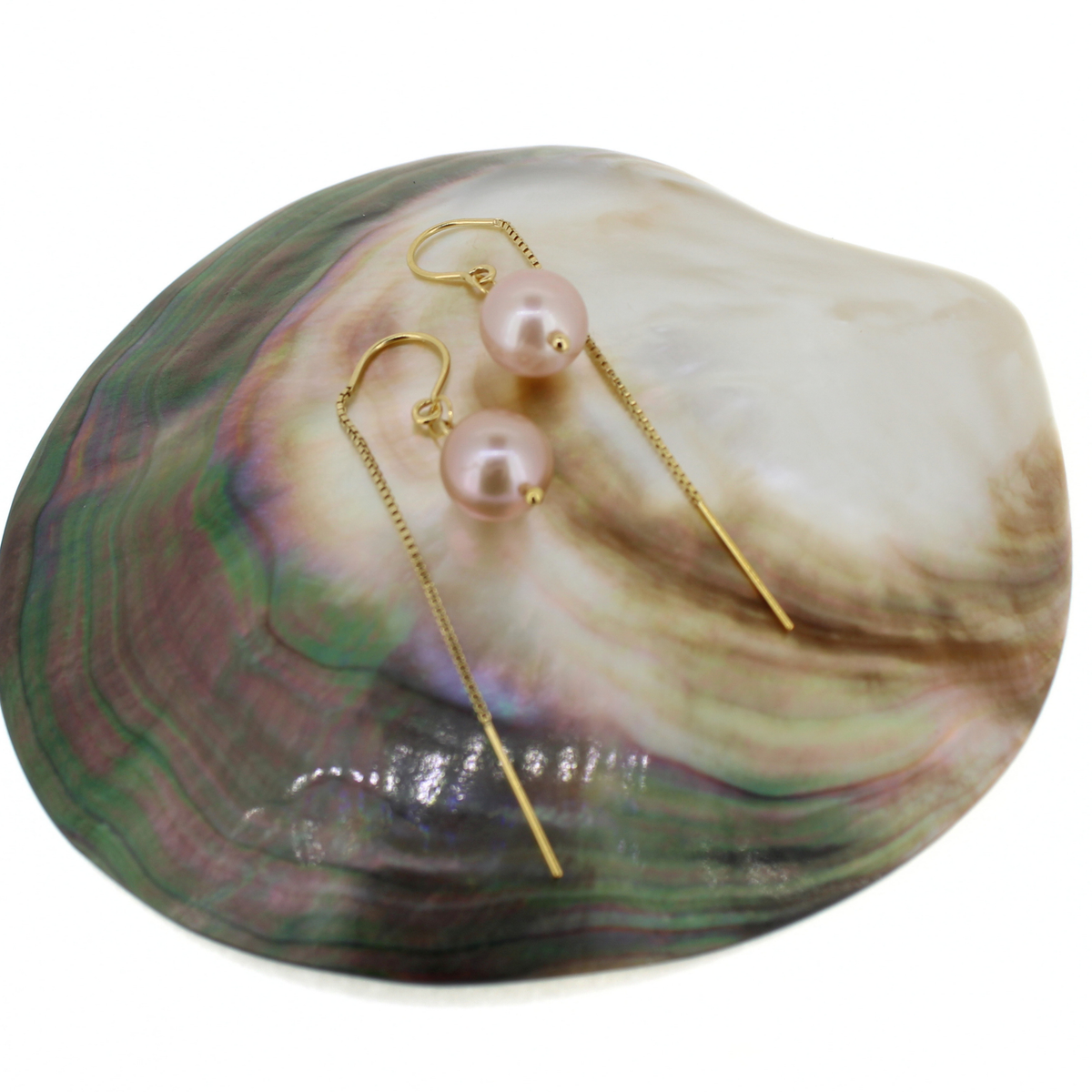 A pair of gold drop earrings with light pink freshwater pearls, displayed on a large iridescent abalone shell against a white background.