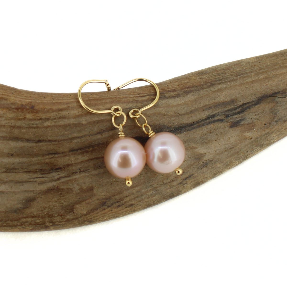 A pair of gold drop earrings with round light pink freshwater pearls, displayed on a curved piece of natural driftwood against a white background.