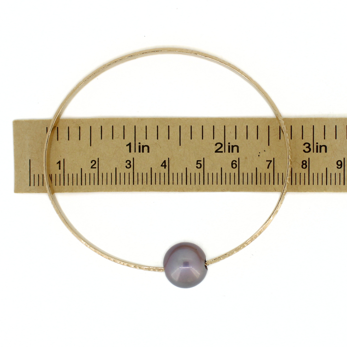 A thin gold wire bracelet with a single round purple pearl bead, shown next to a paper ruler for scale on a white background.