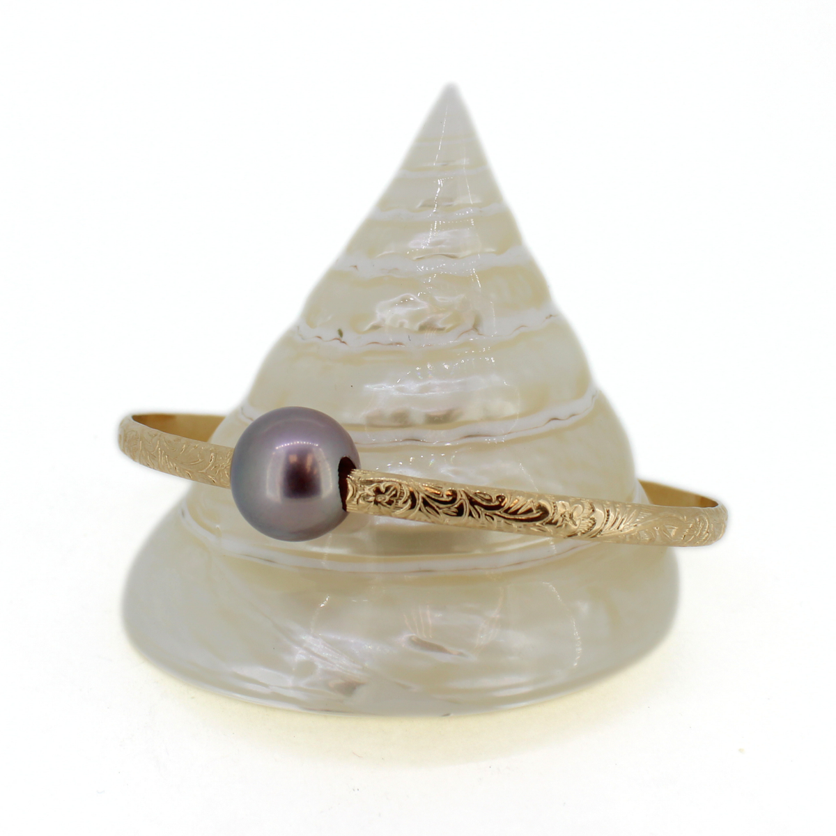 A gold bangle bracelet with an engraved floral pattern and a single round dark purple pearl, displayed on a white seashell stand against a white background.