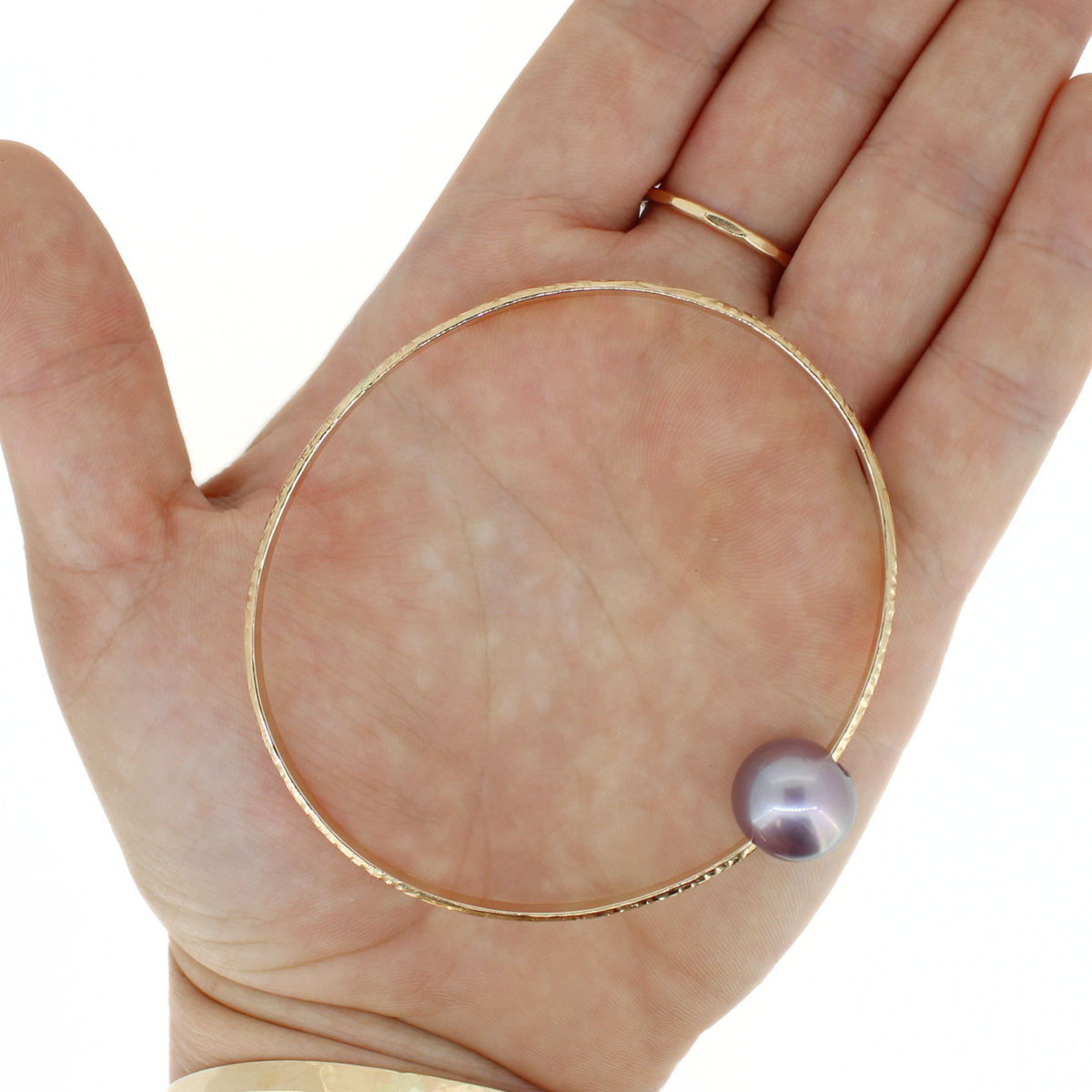 A gold bangle bracelet with a single round purple pearl, shown resting on an open hand. Set against a clean white background.