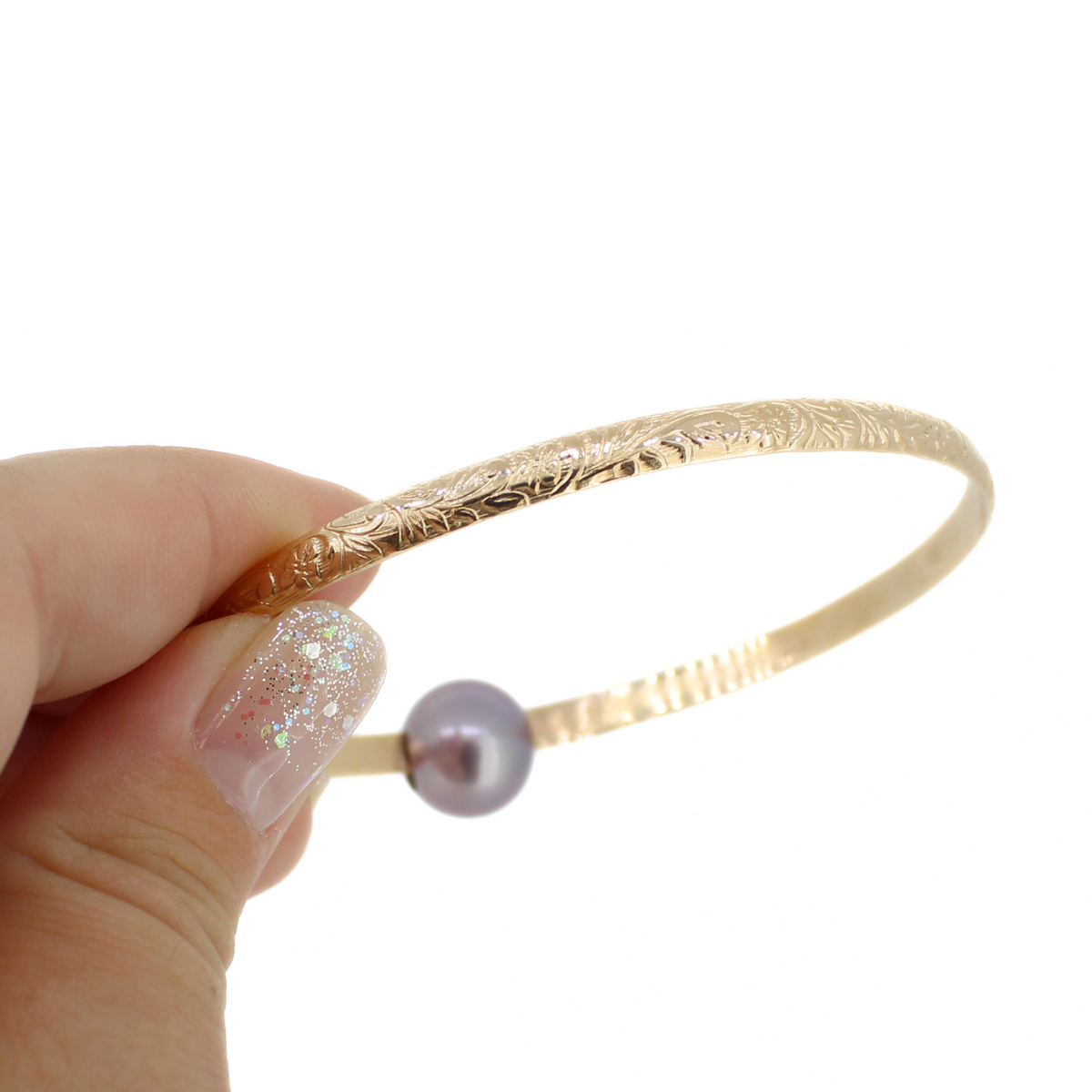 A hand with glittery nail polish holds a delicate gold bangle bracelet featuring an engraved floral pattern and a small round purple pearl bead, set against a plain white background.