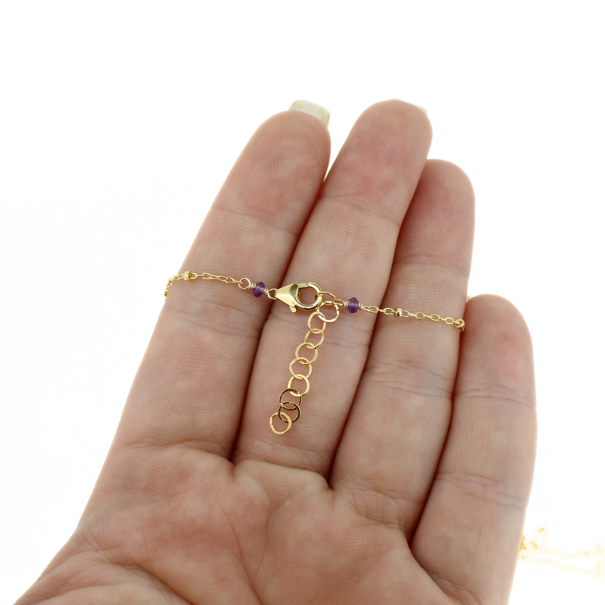 A delicate gold chain necklace with a lobster clasp and purple accent beads, shown resting on an open hand against a white background.