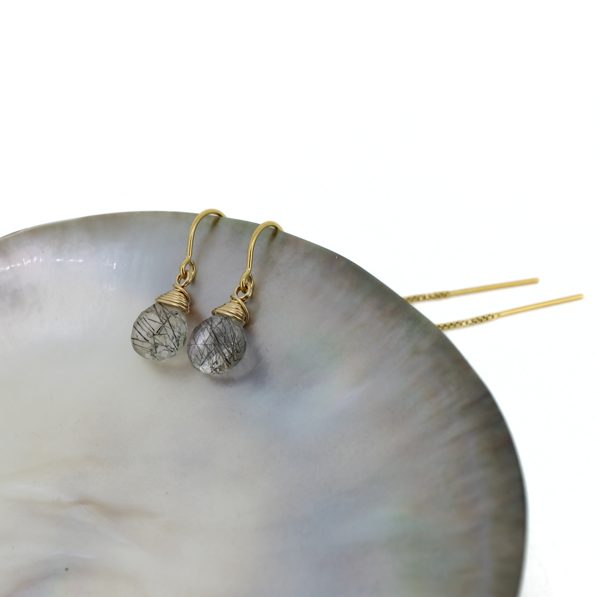 A pair of gold-tone drop earrings with teardrop-shaped gray and black striped stone beads, displayed on a curved iridescent shell dish against a white background.