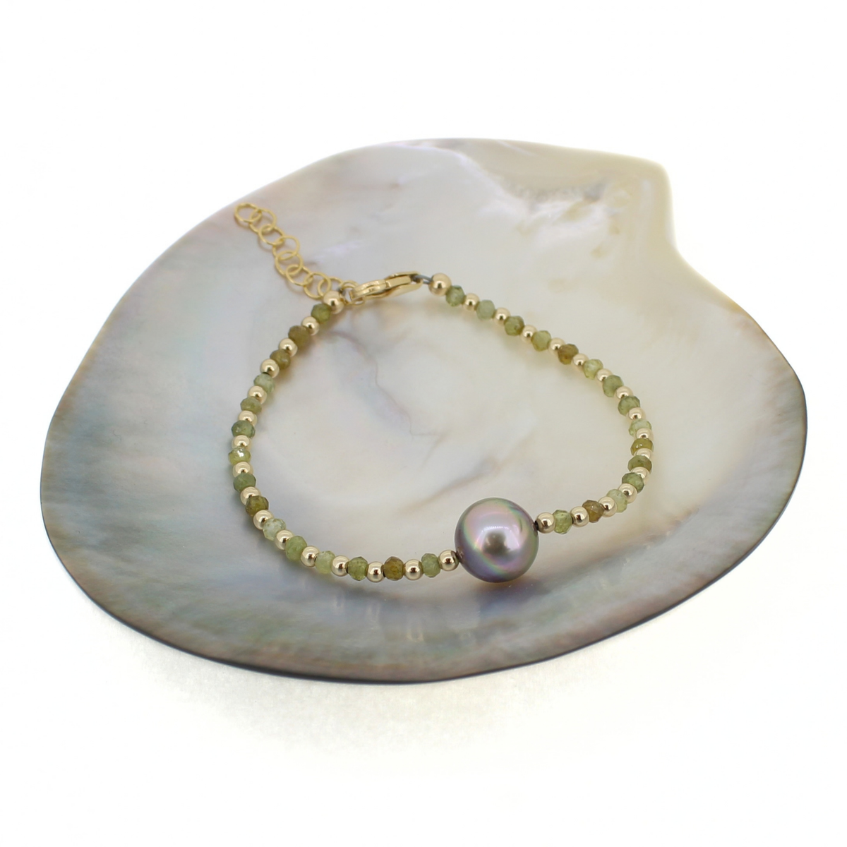 Gold beaded bracelet featuring faceted green garnet gemstones and a gray Tahitian pearl at the center. Displayed on a mother of pearl shell.