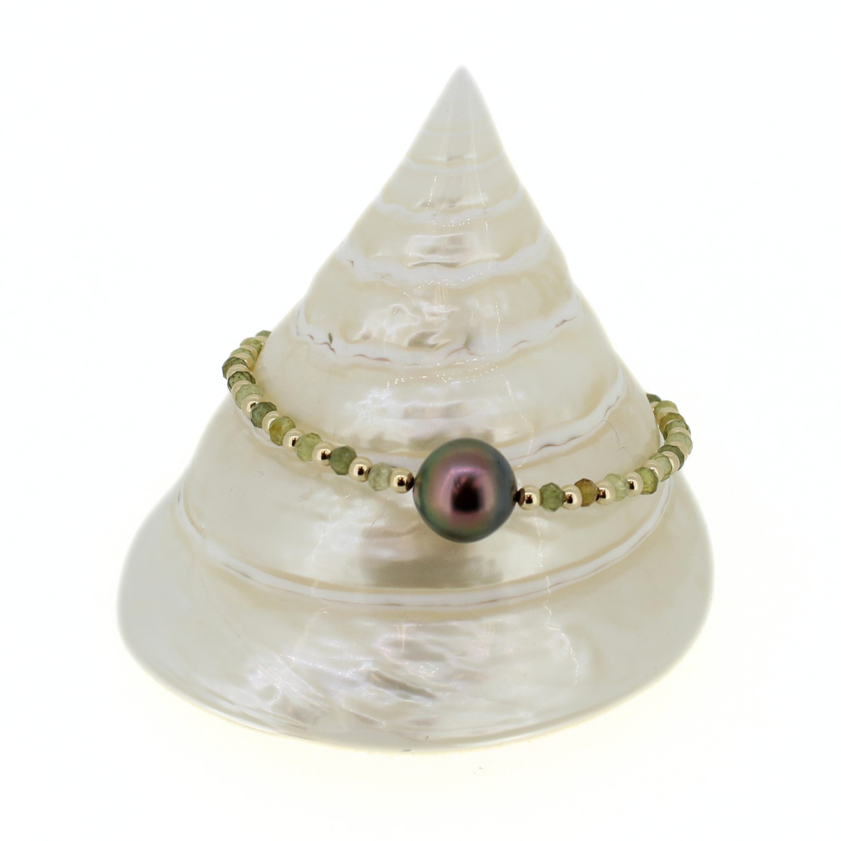 A beaded bracelet with a central iridescent dark pearl and small gold spacer beads, displayed on a white seashell stand against a white background.