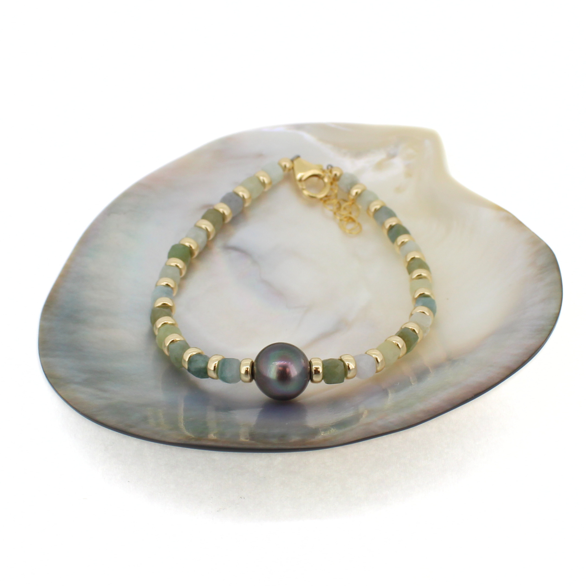 A beaded bracelet with gold-toned spacer beads, light to medium green cube shaped jade beads, and a large dark gray Tahitian pearl centerpiece, displayed on a white shell dish against a white background.