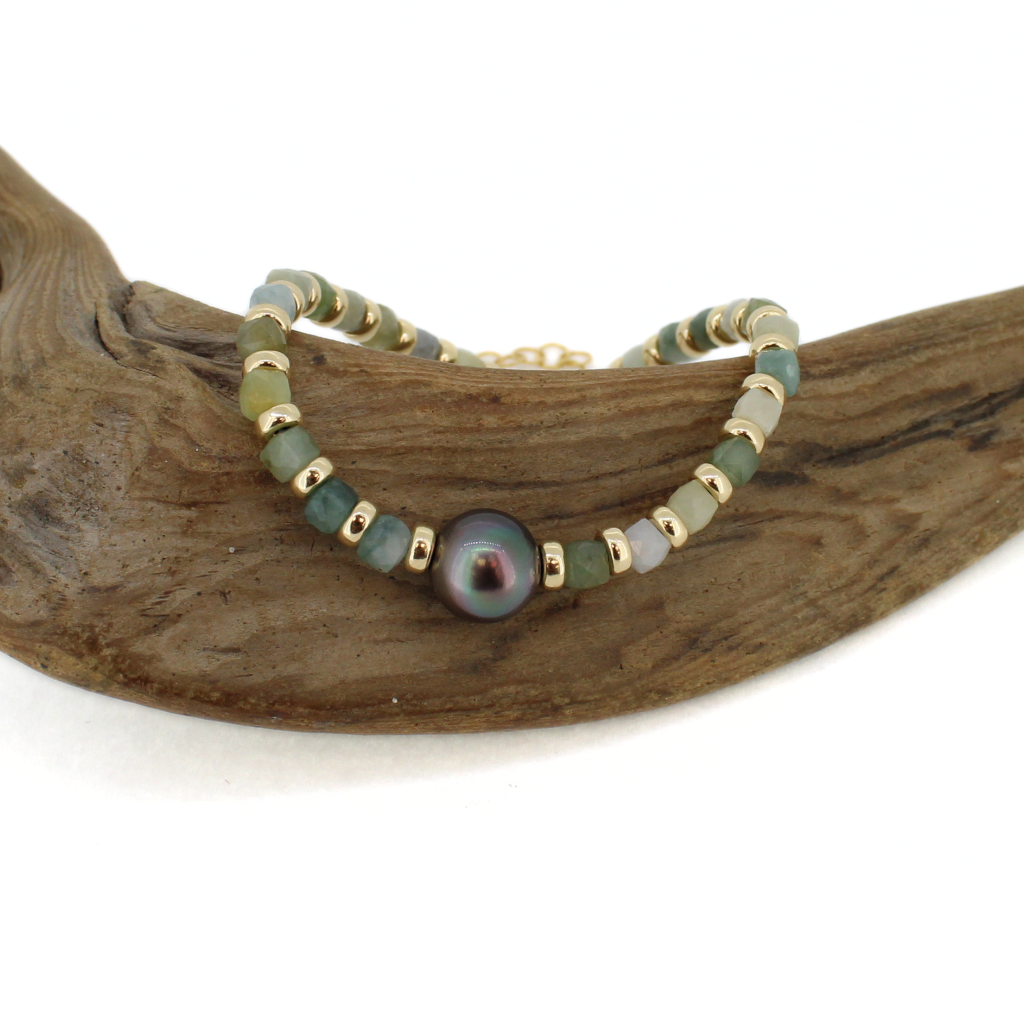 A beaded bracelet with green jade gemstone beads, gold spacer beads, and a central iridescent dark Tahitian pearl, displayed on a curved piece of driftwood against a white background.