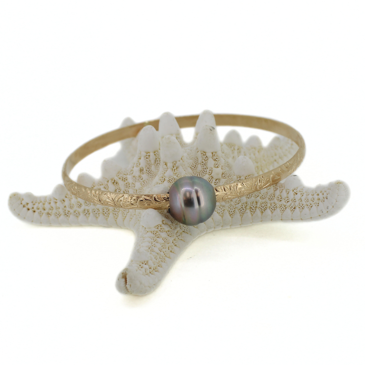 A gold bangle bracelet with a single iridescent pearl bead, displayed on a white starfish prop against a white background.