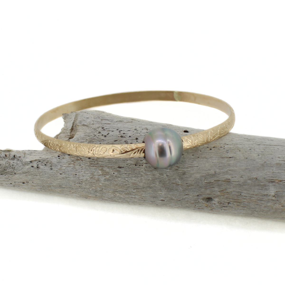 A delicate gold bangle bracelet with a floral textured band and a single iridescent grey pearl, resting on a piece of weathered driftwood against a white background.