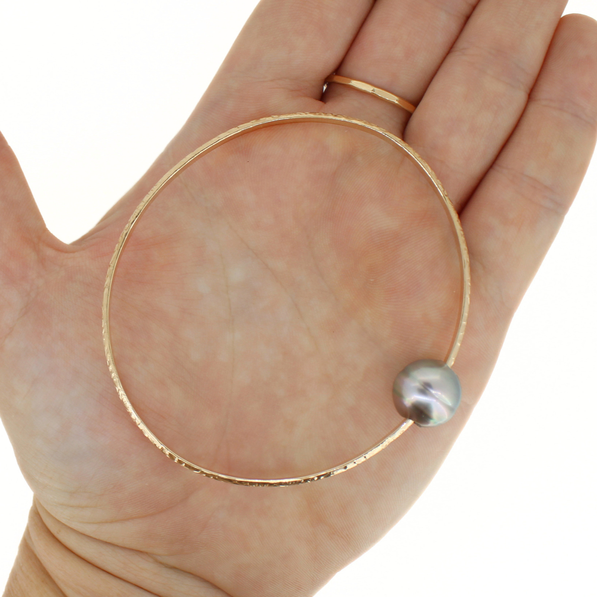 A textured gold bangle bracelet with a single round gray pearl bead, shown resting on an open hand.