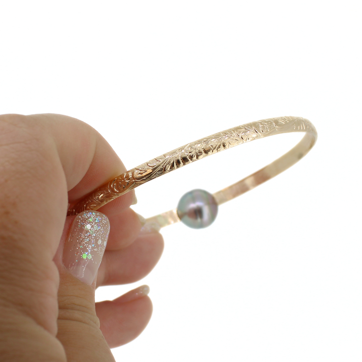 A hand holds a gold bangle bracelet featuring an engraved floral pattern and a small iridescent pearl, set against a plain white background.