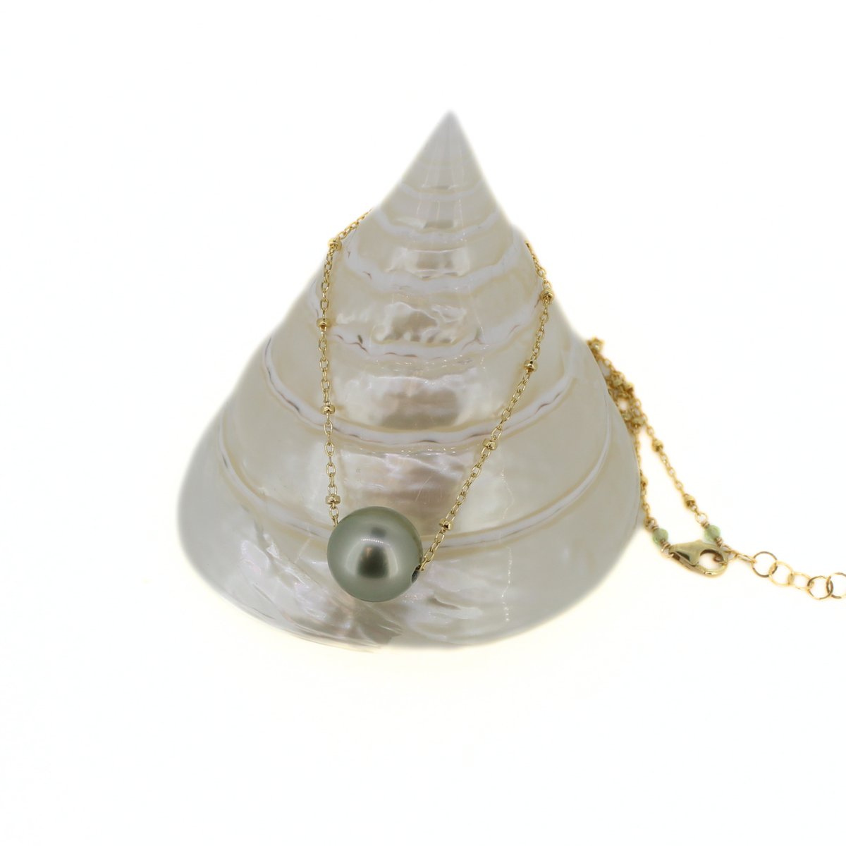 A gold chain necklace with a single round green pearl pendant, draped over a white seashell display on a white background.