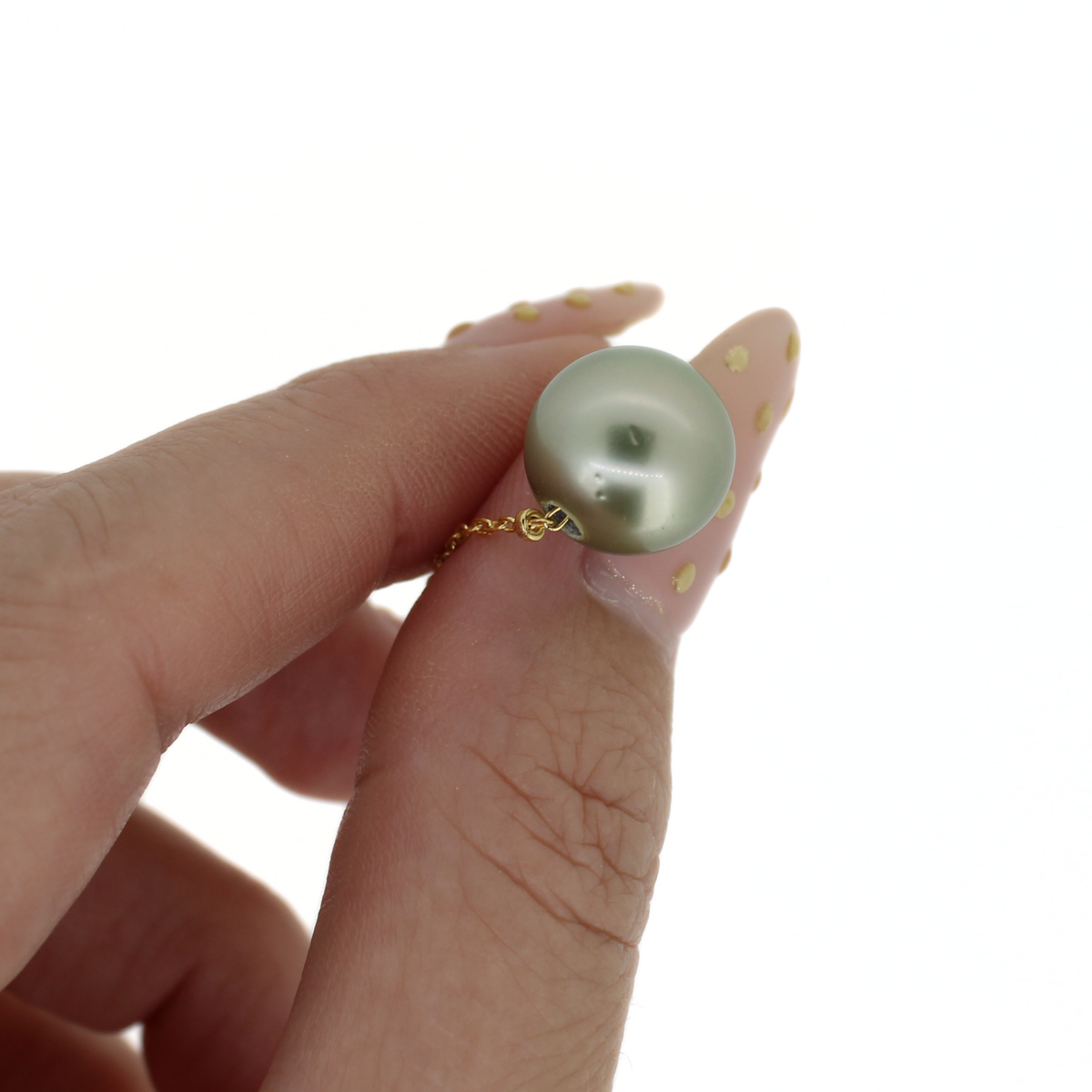 A close-up of a hand with long, manicured nails holding a gold necklace with a round, lustrous pale green pearl pendant against a solid white background.
