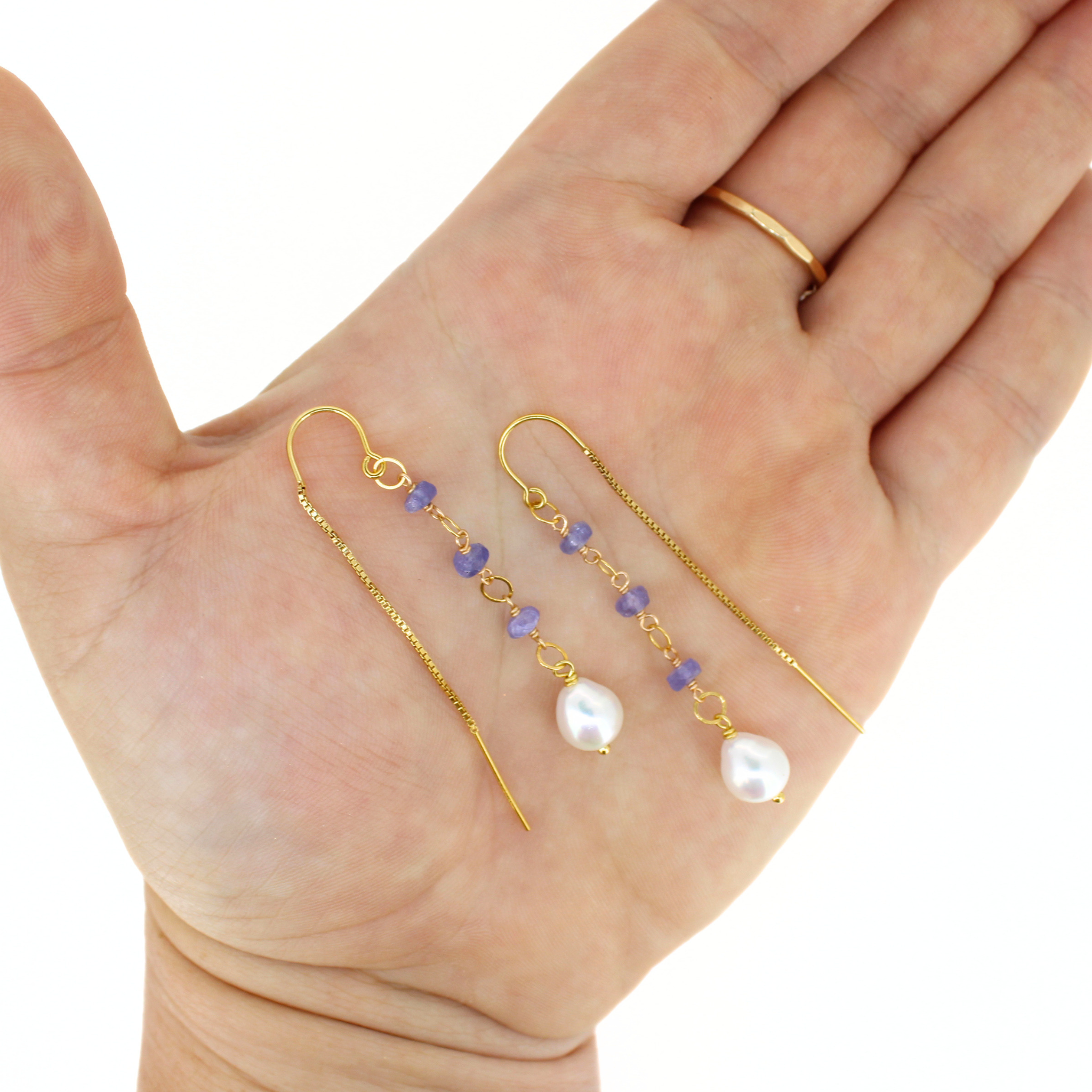 A pair of gold drop earrings with small purple beads and white pearl-like drops, displayed on an open hand against a white background.