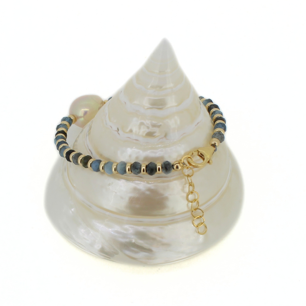 A beaded bracelet with dark gray and light blue round beads, accented by small gold spacer beads and a gold lobster clasp, displayed on a white seashell jewelry stand against a white background.