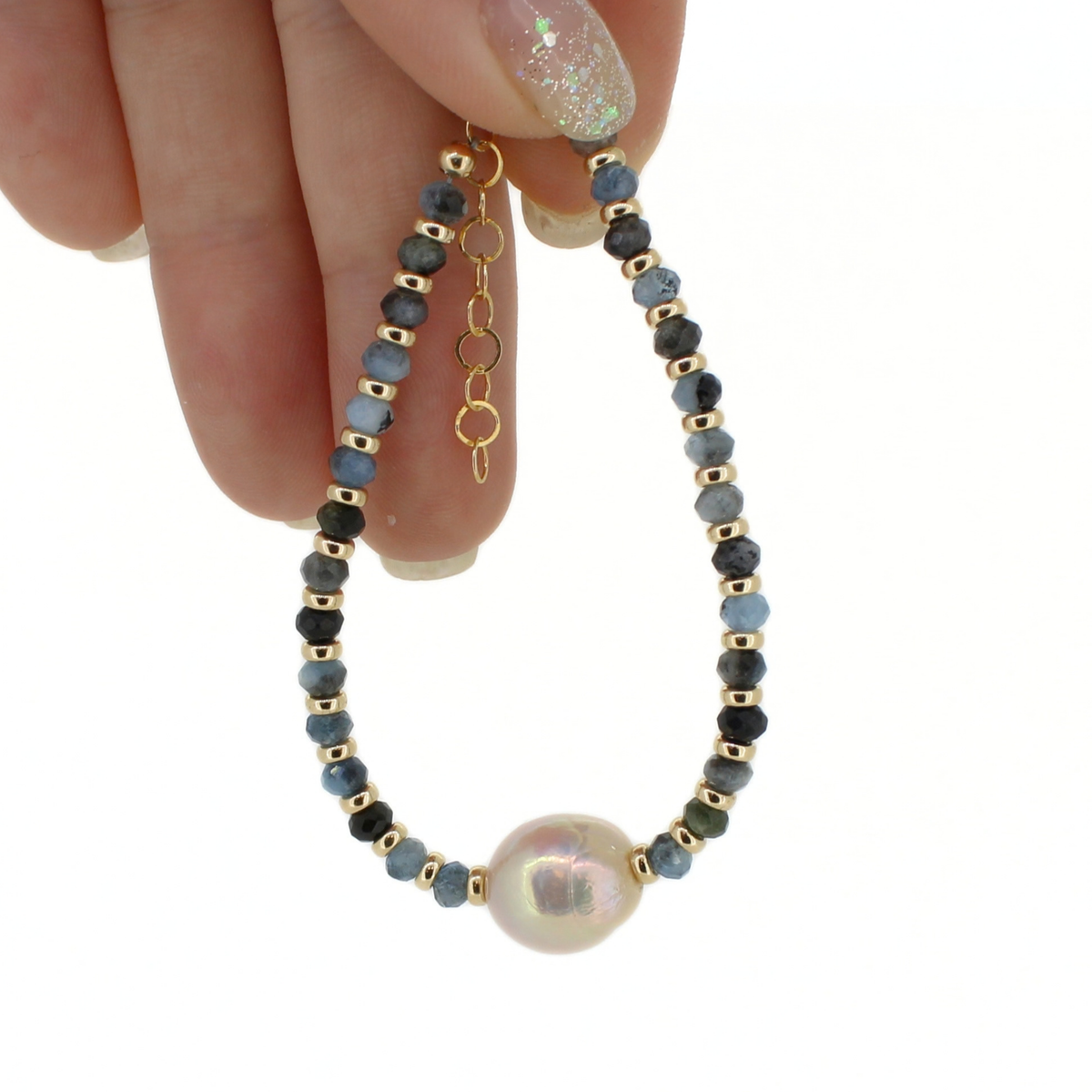 A delicate beaded bracelet with faceted blue-gray beads, small gold spacer beads, and a large iridescent pearl at the center, held by a hand against a white background.