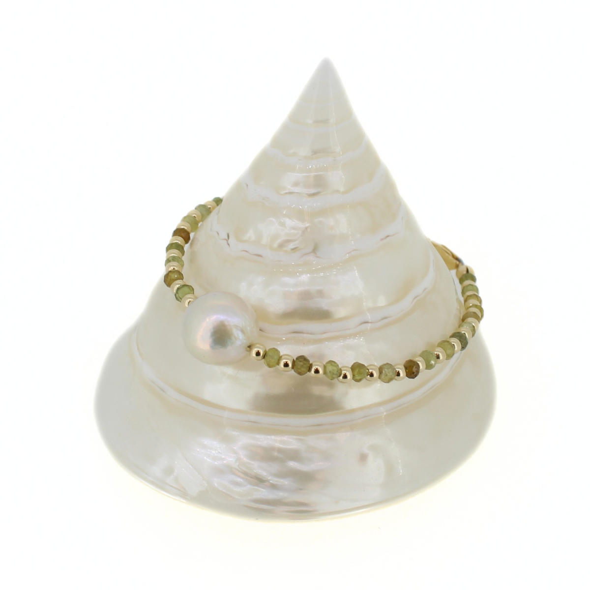 A gold beaded bracelet with a single white pearl bead, displayed on a white mother-of-pearl seashell jewelry stand against a white background.