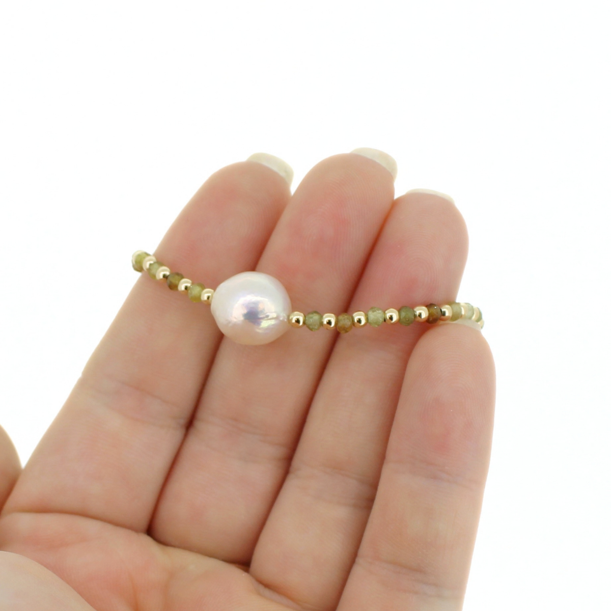 A delicate beaded bracelet with a large white pearl and small gold beads, held on a person's fingers against a plain white background.