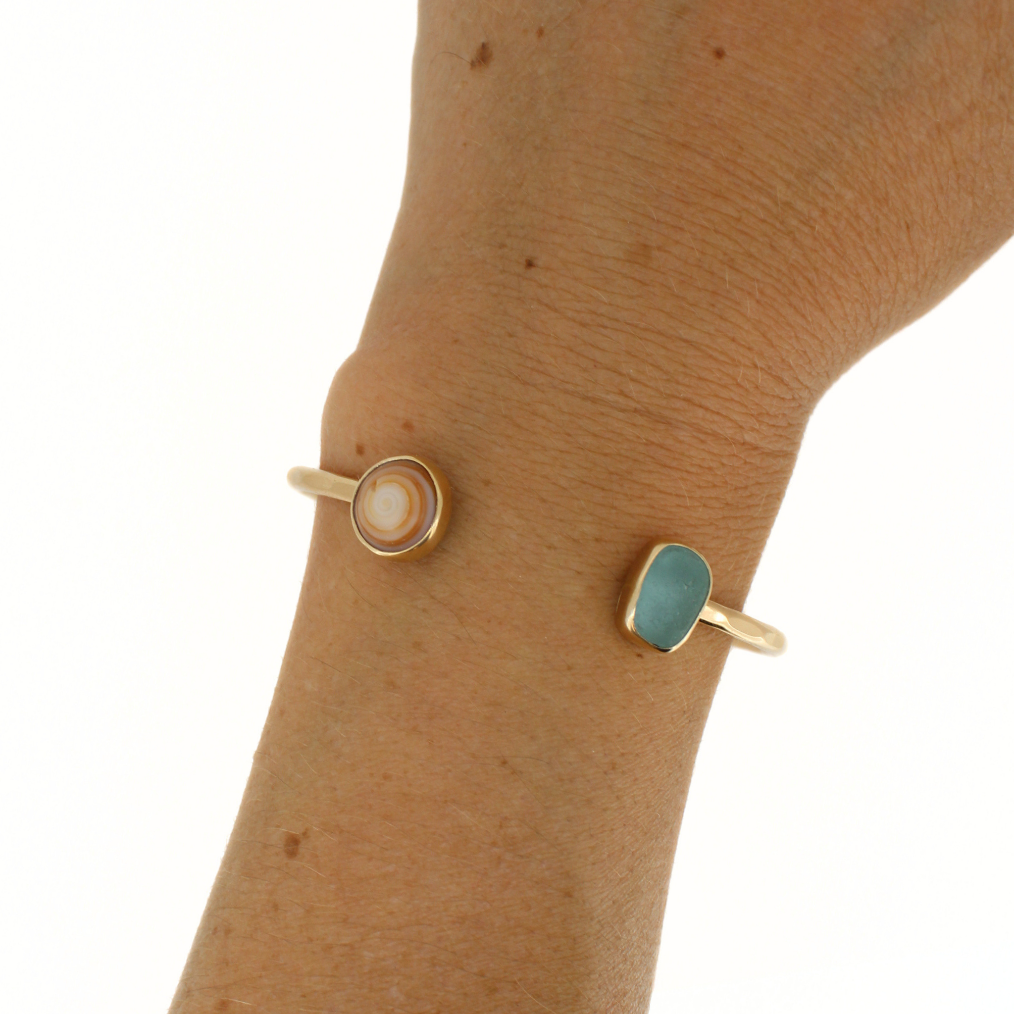 Gold cuff bracelet on a wrist featuring a brown Hawaiian puka shell and genuine blue sea glass.