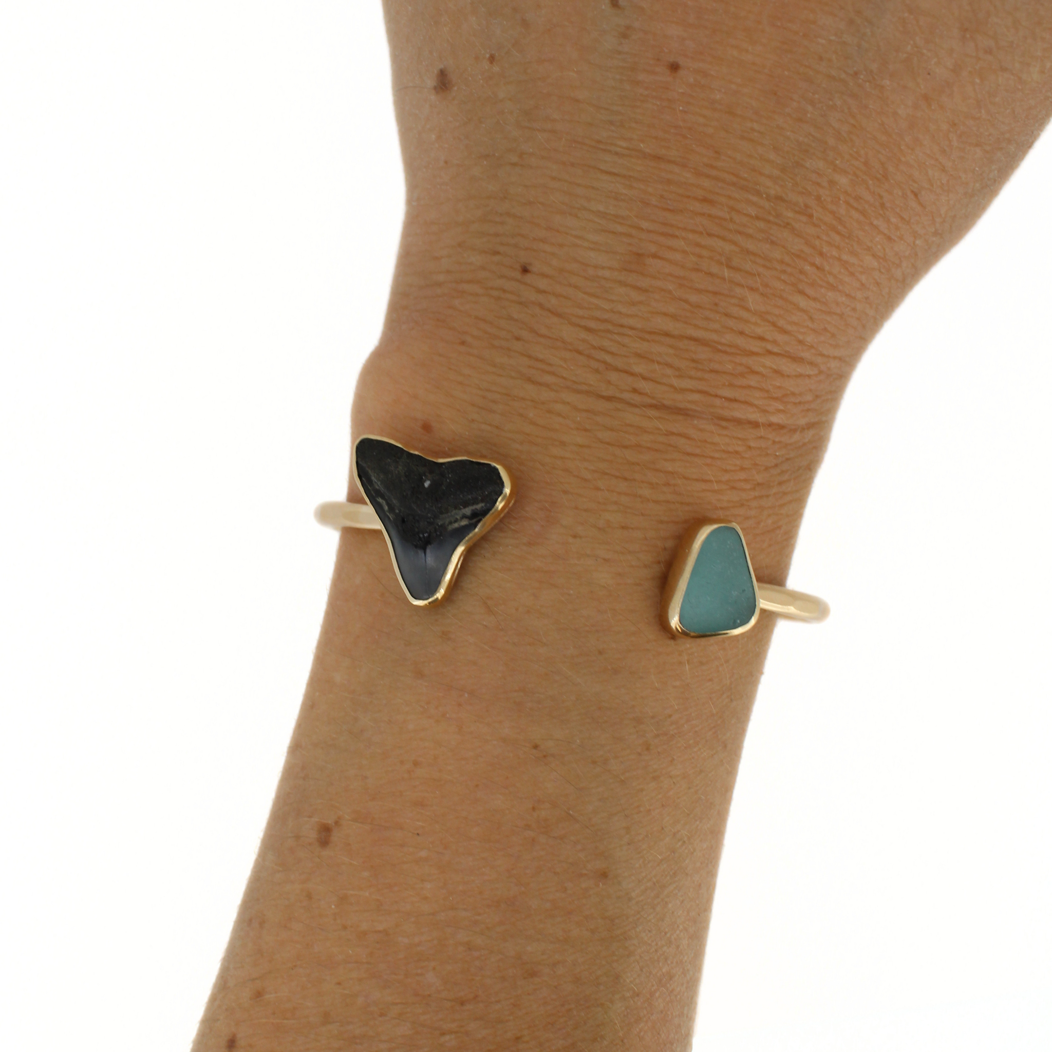 Gold cuff bracelet on wrist with black shark tooth and teal sea-glass triangle
