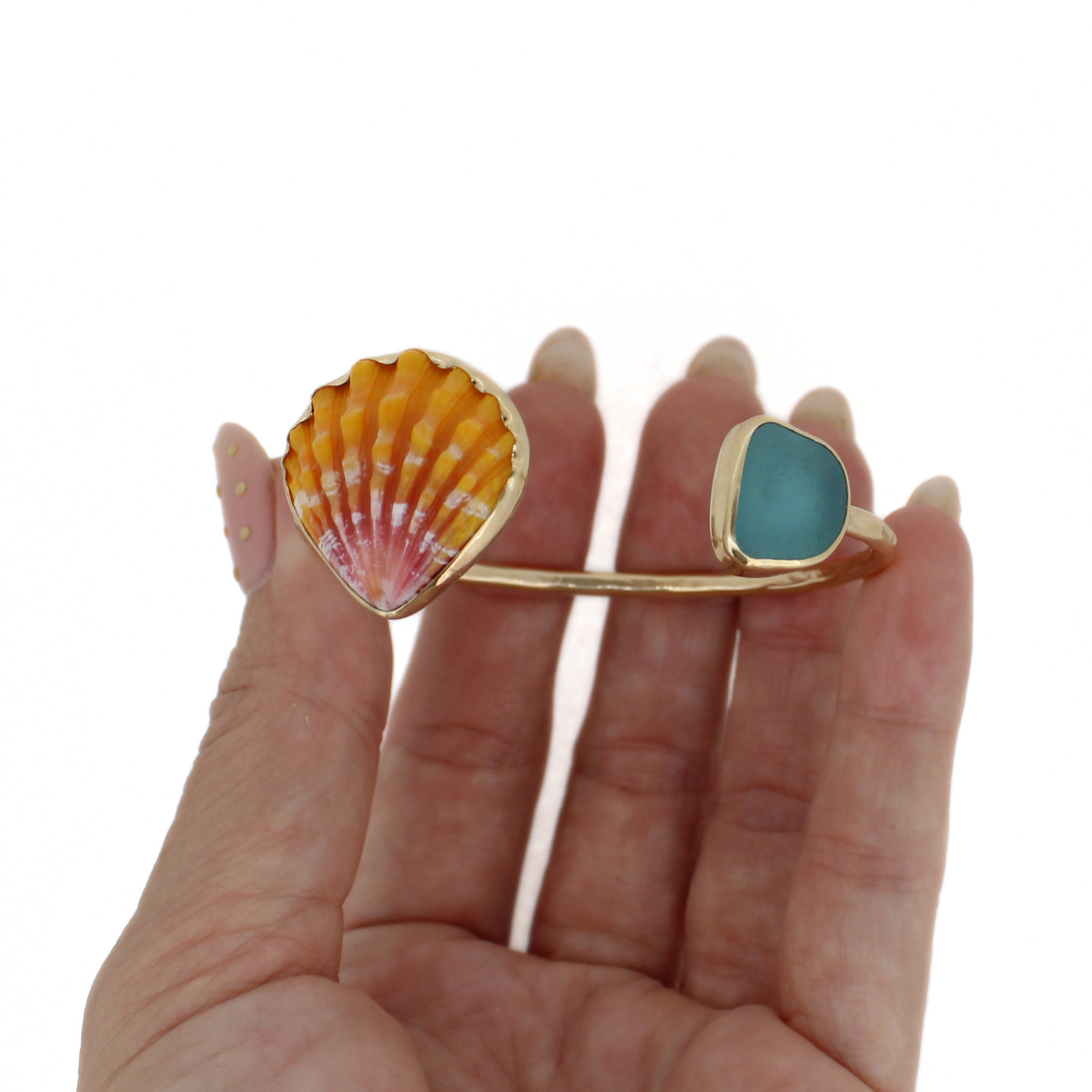 Gold cuff bracelet held in hand: teal sea-glass square and orange sunrise shell