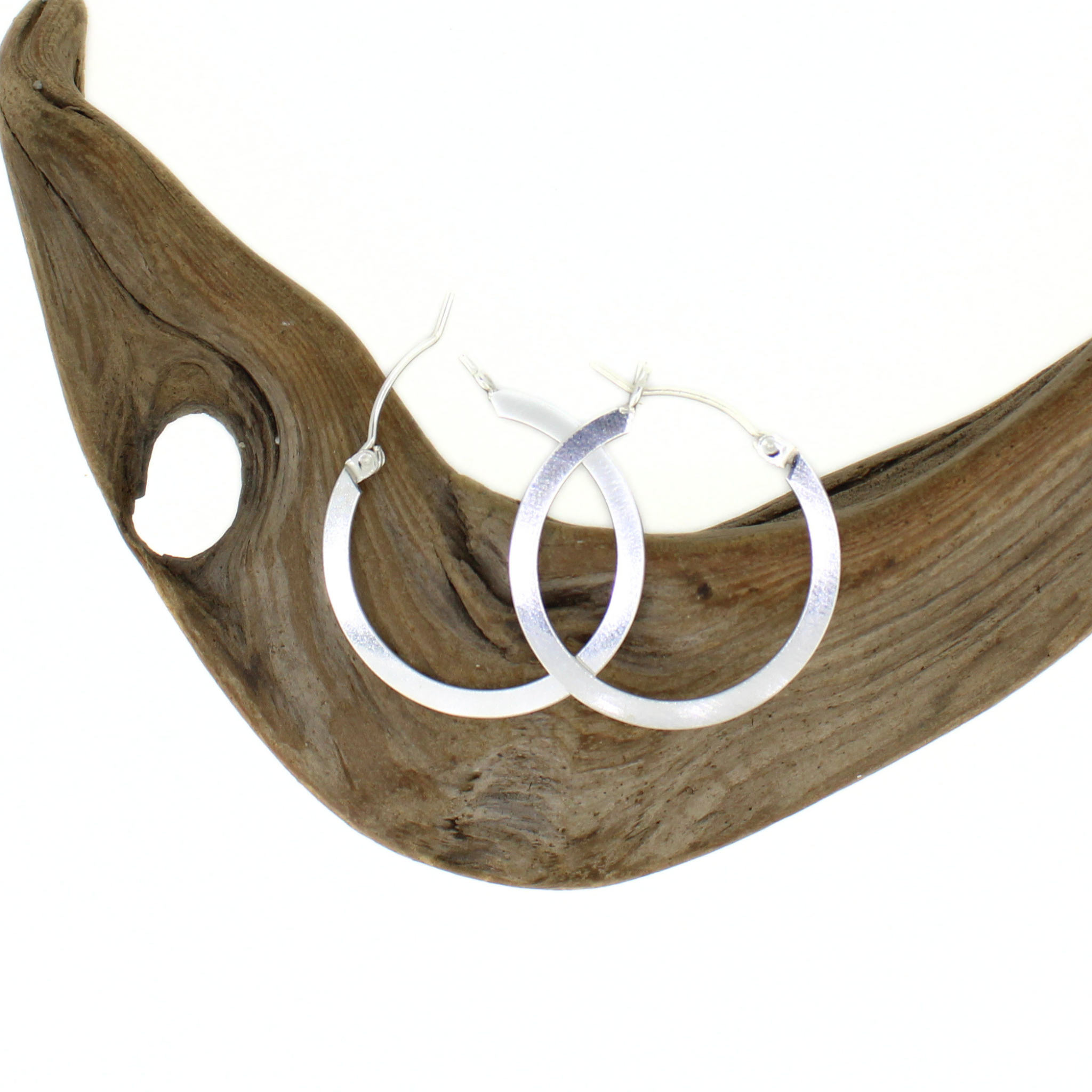 A pair of silver hoop earrings with a shiny finish, displayed on a curved piece of natural driftwood against a solid white background.
