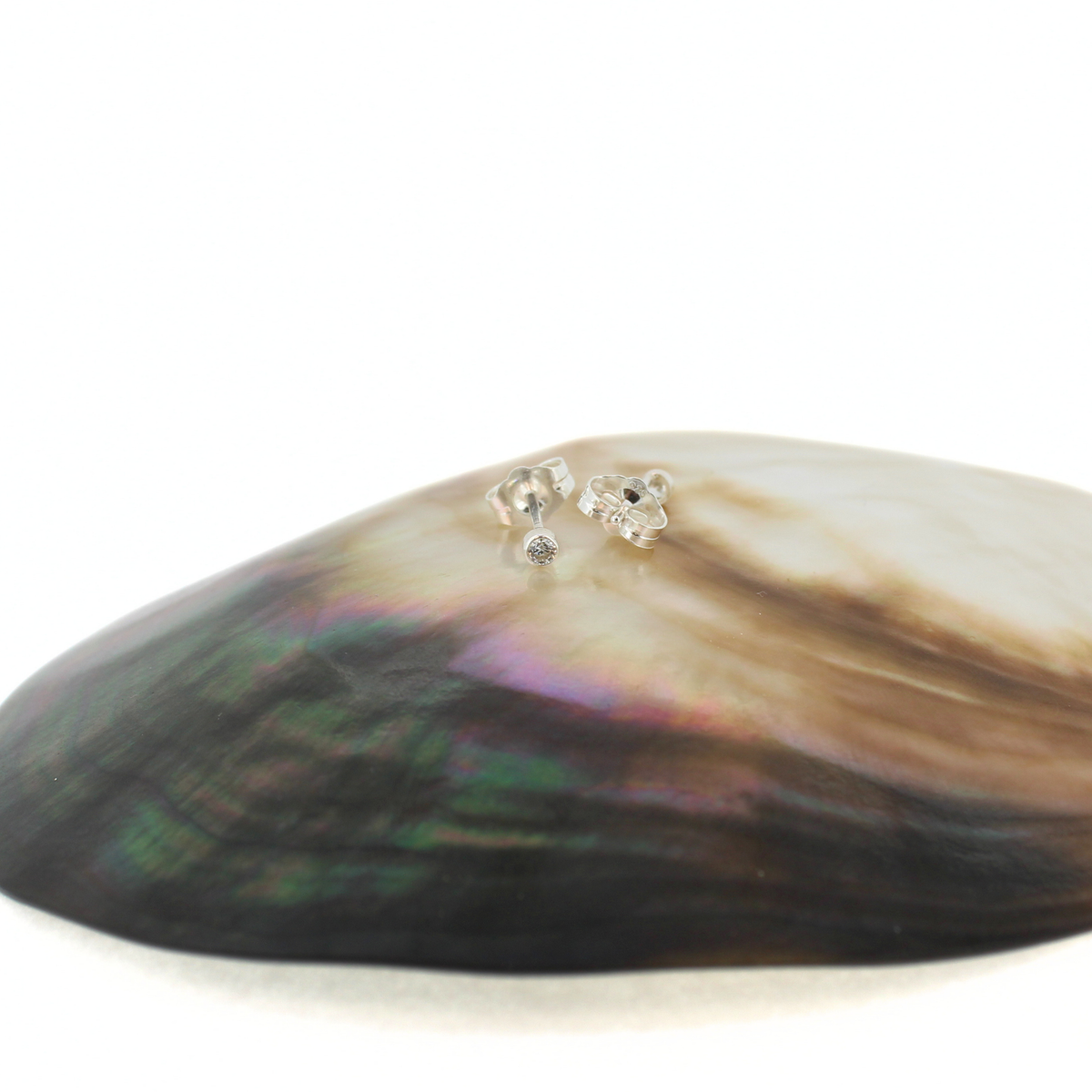 A pair of small silver stud earrings with clear stones, displayed on top of an iridescent abalone shell against a white background.
