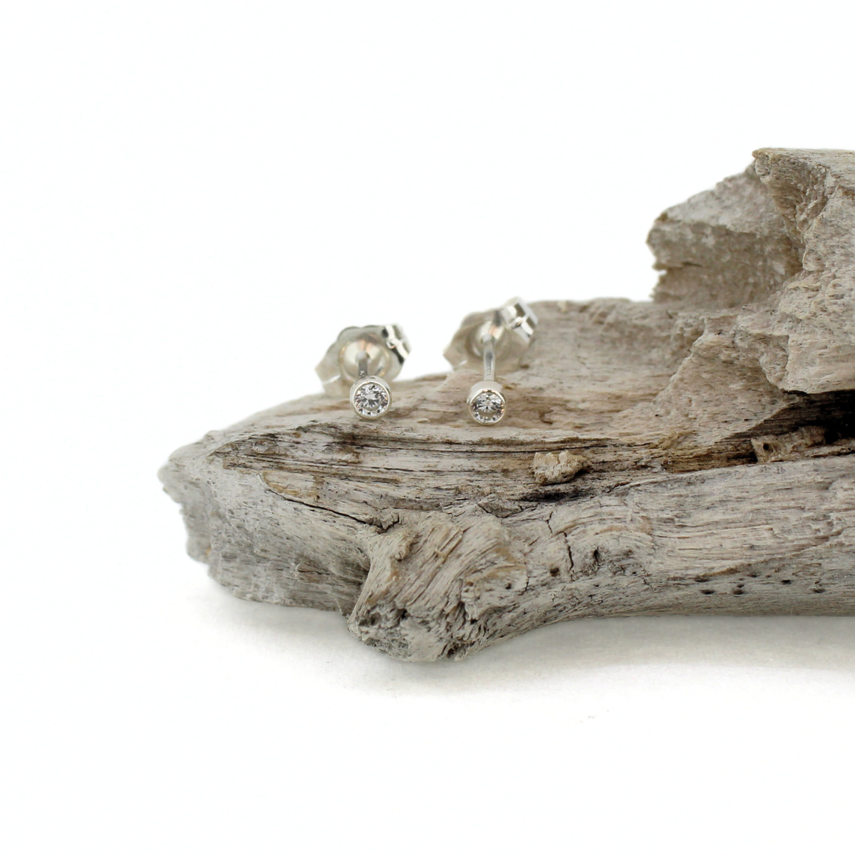 A pair of silver stud earrings with clear round gemstones, displayed on a piece of weathered driftwood against a white background.