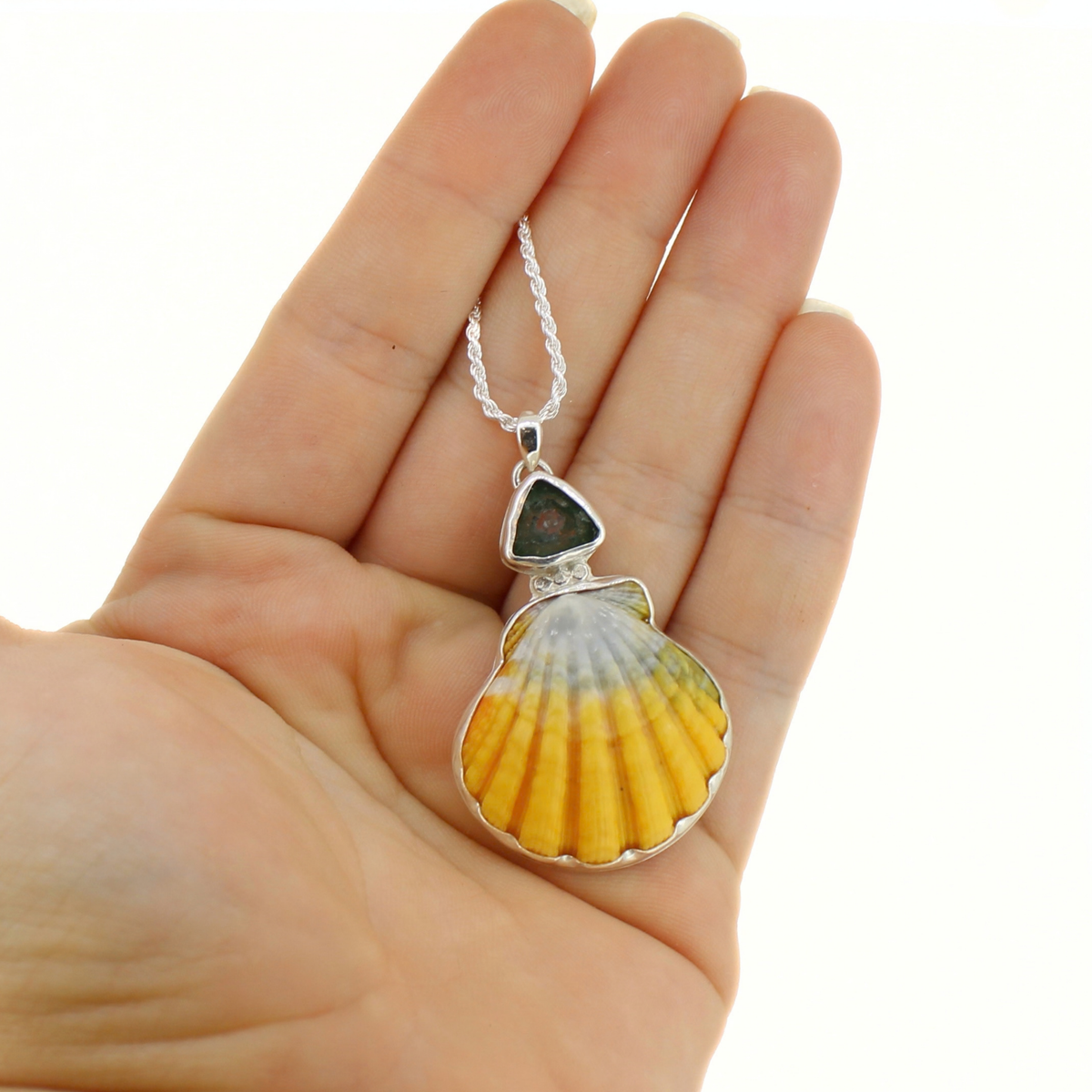 A silver scallop shell pendant necklace with a yellow and white gradient shell design, held in a person's hand against a white background.