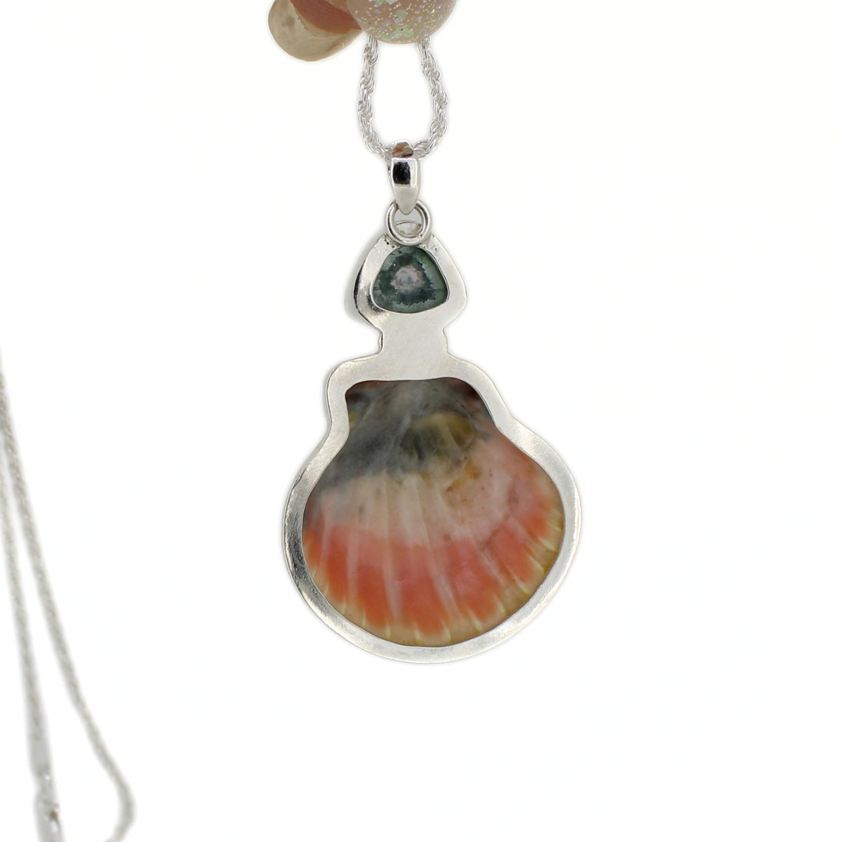 The backside of a silver pendant necklace featuring a scallop shell inlay with pink, orange, and cream tones, accented by a small blue stone at the top, shown on a white background.