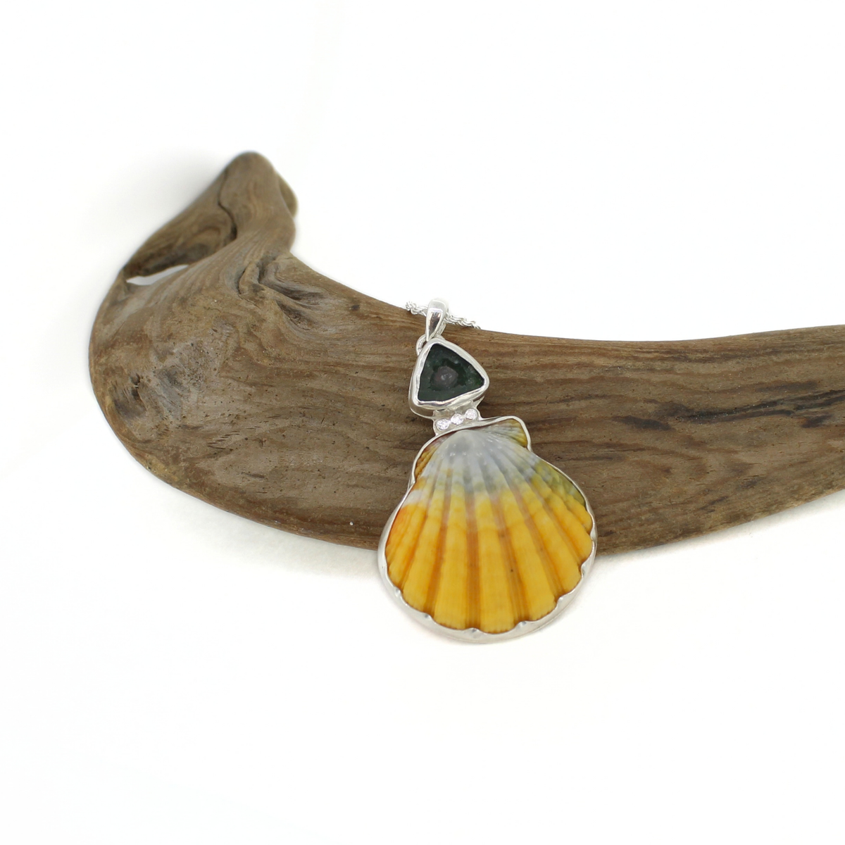 A silver shell pendant featuring a yellow and gray sunrise shell design with a small dark blue teardrop stone at the top, displayed on a piece of driftwood against a white background.