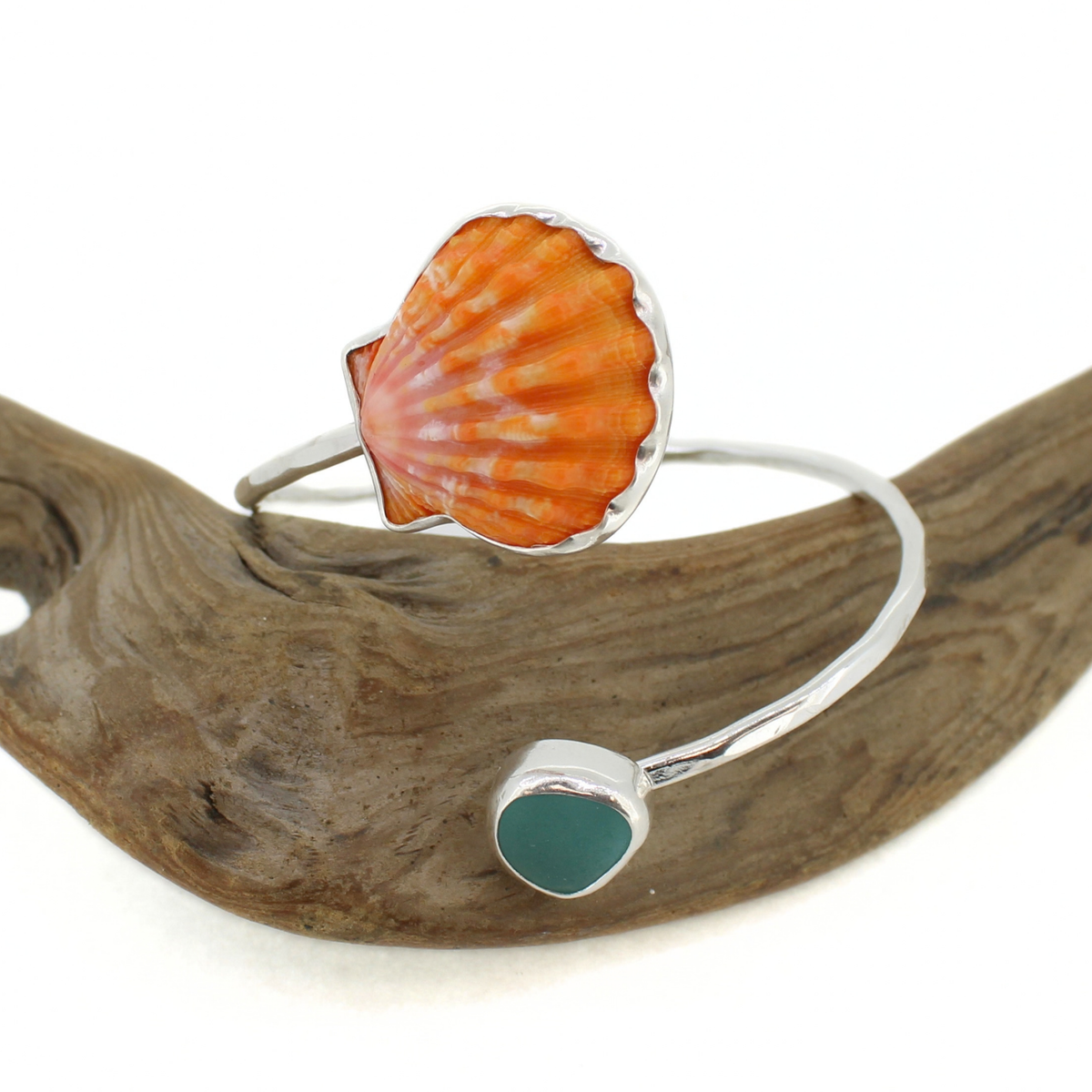 A silver cuff bracelet featuring a scallop shell in orange and white on one end and a small blue sea glass on the other, displayed on a piece of driftwood against a white background.