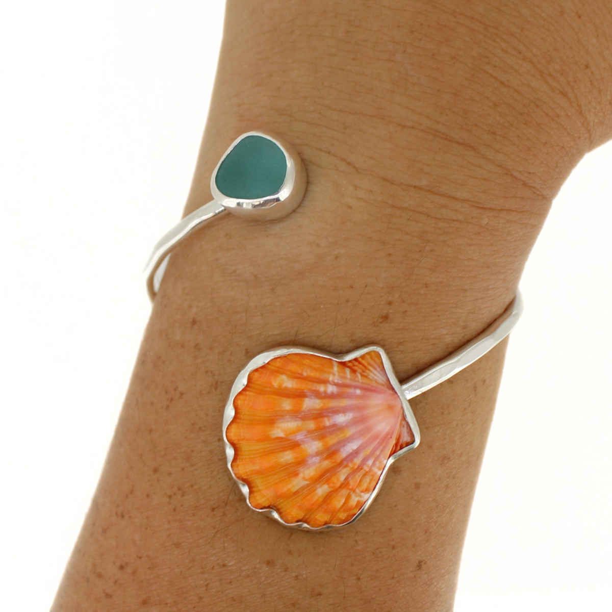 A sterling silver open cuff bracelet with a smooth sea glass and orange and pink sunrise seashell, shown on a person's wrist.