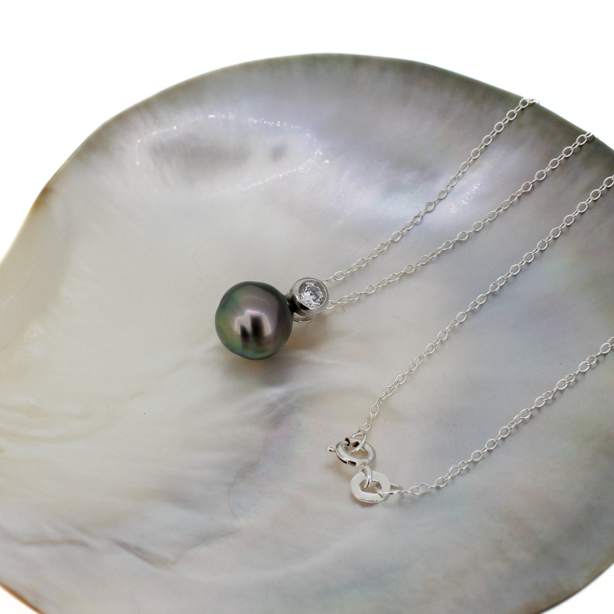 A silver chain necklace with a dark gray-green Tahitian pearl pendant and a small clear accent stone, displayed on a white mother-of-pearl shell dish against a white background.