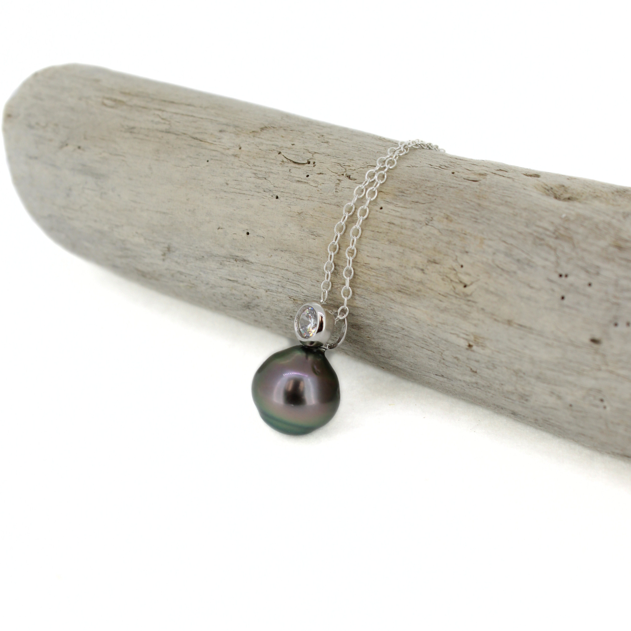 A silver chain necklace with a dark, iridescent baroque pearl pendant and a small clear stone accent, displayed on a piece of driftwood against a white background.