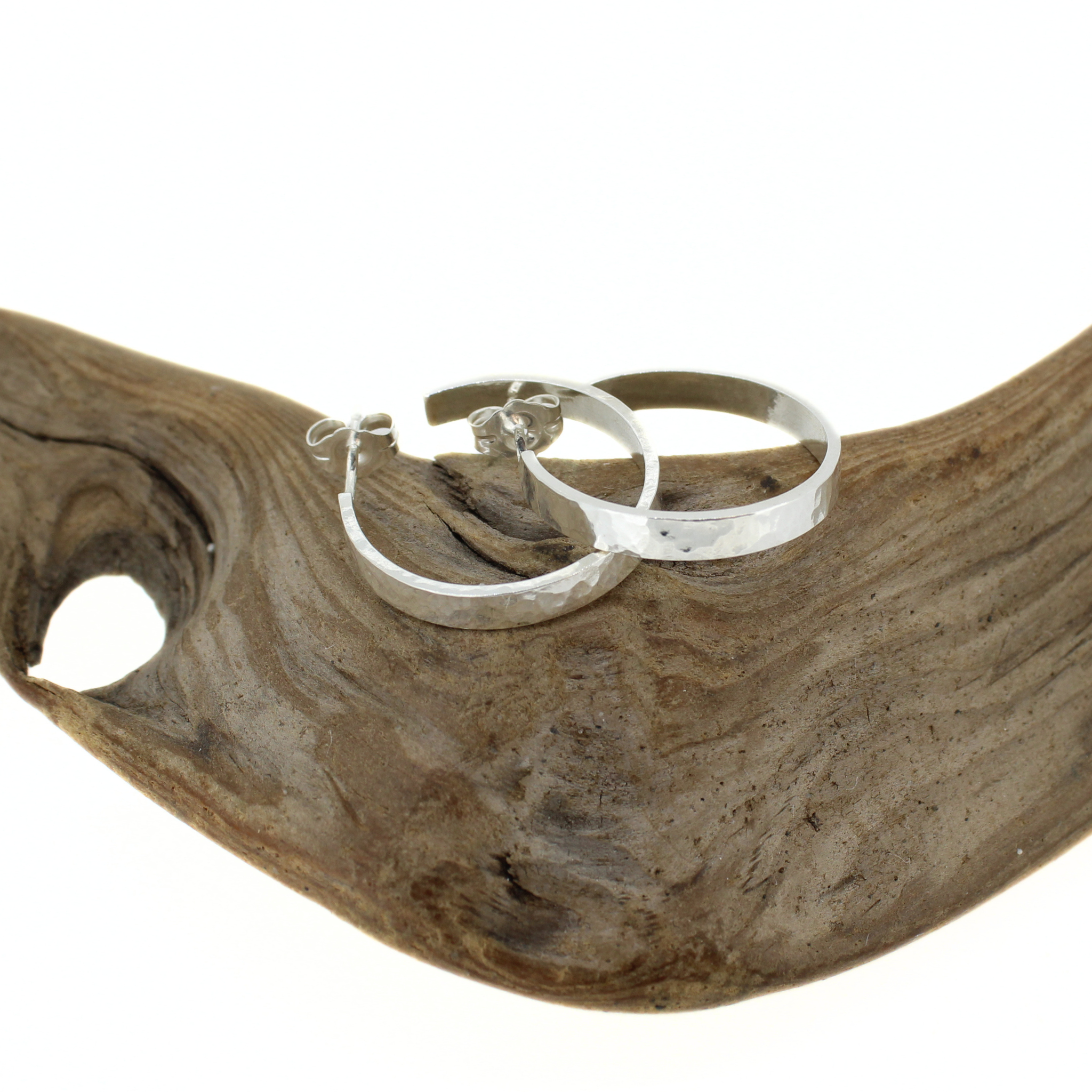 A pair of silver hoop earrings with a hammered texture, displayed on a curved piece of natural driftwood against a white background. Handmade by Brianne & Co.