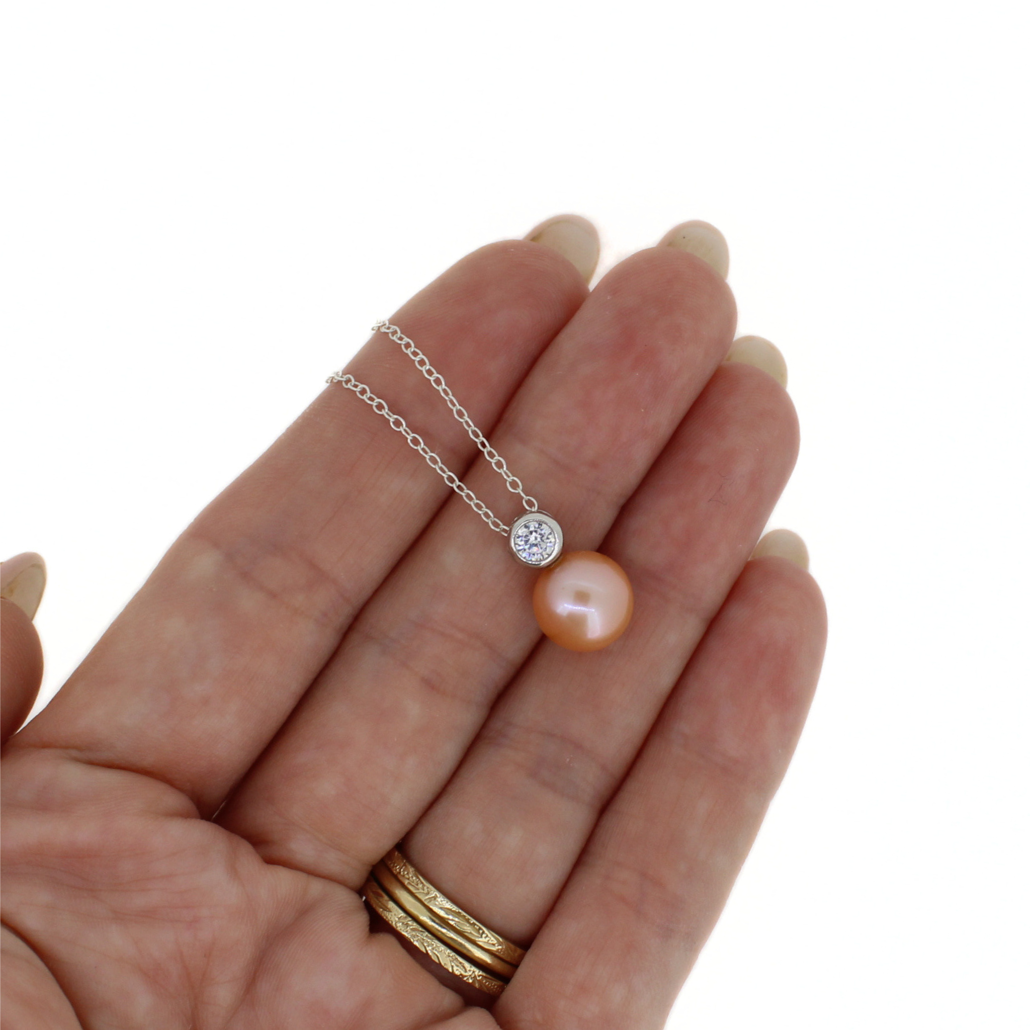 A delicate silver chain necklace with a single peach-pink pearl pendant and a small round clear stone setting, shown resting on an open hand against a white background.