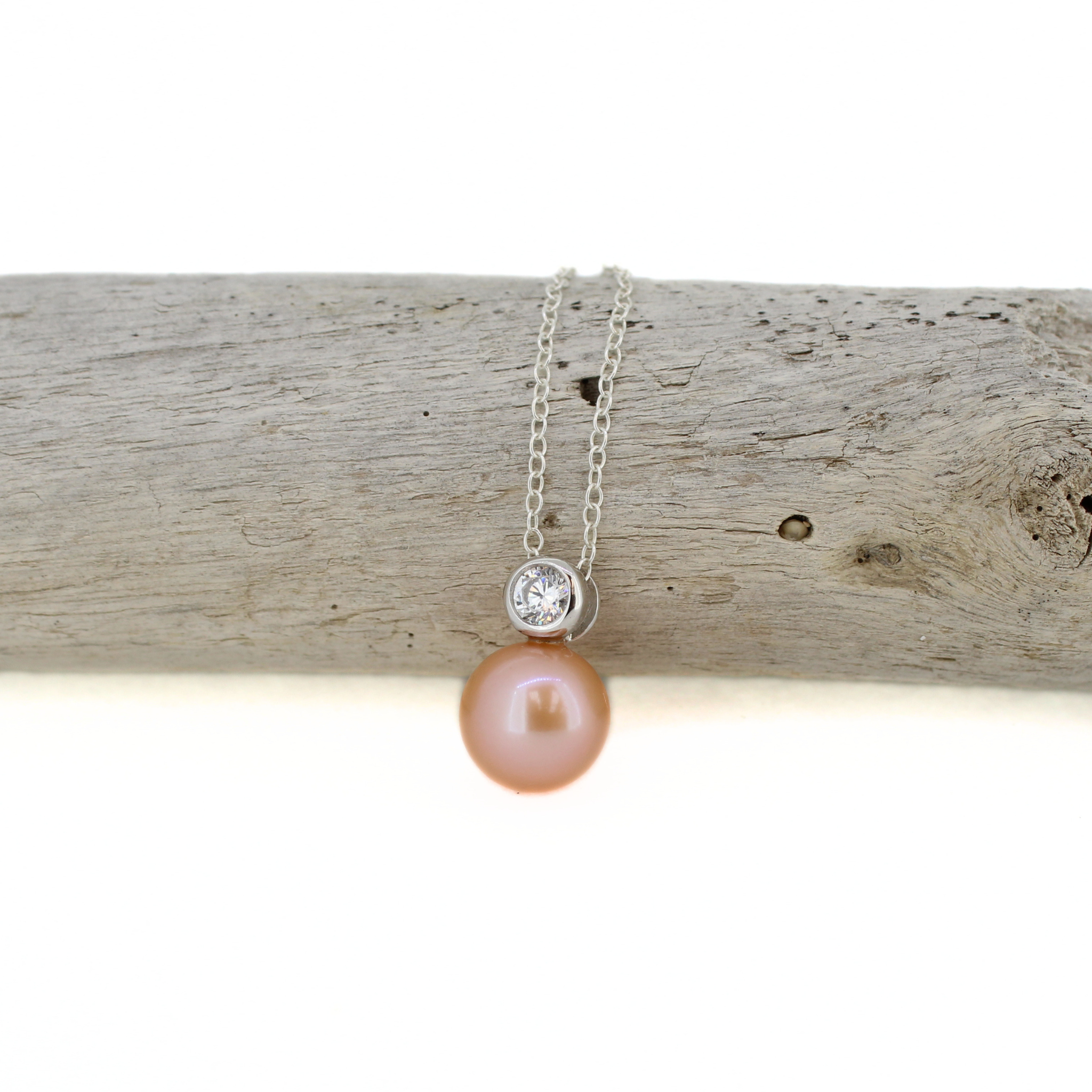 A silver chain necklace featuring a round pink pearl pendant and a small clear round stone in a silver bezel, displayed draped over a piece of weathered driftwood against a white background.