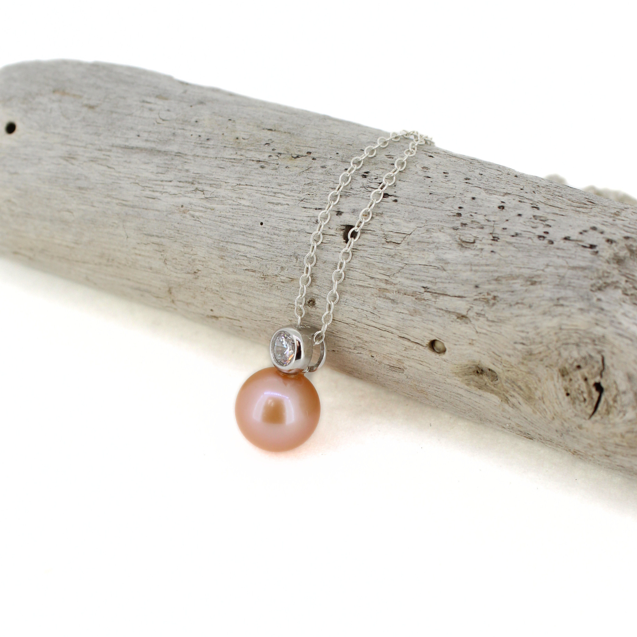 A silver chain necklace with a peach-pink pearl pendant and a small clear stone bezel, displayed on a piece of weathered driftwood against a white background.