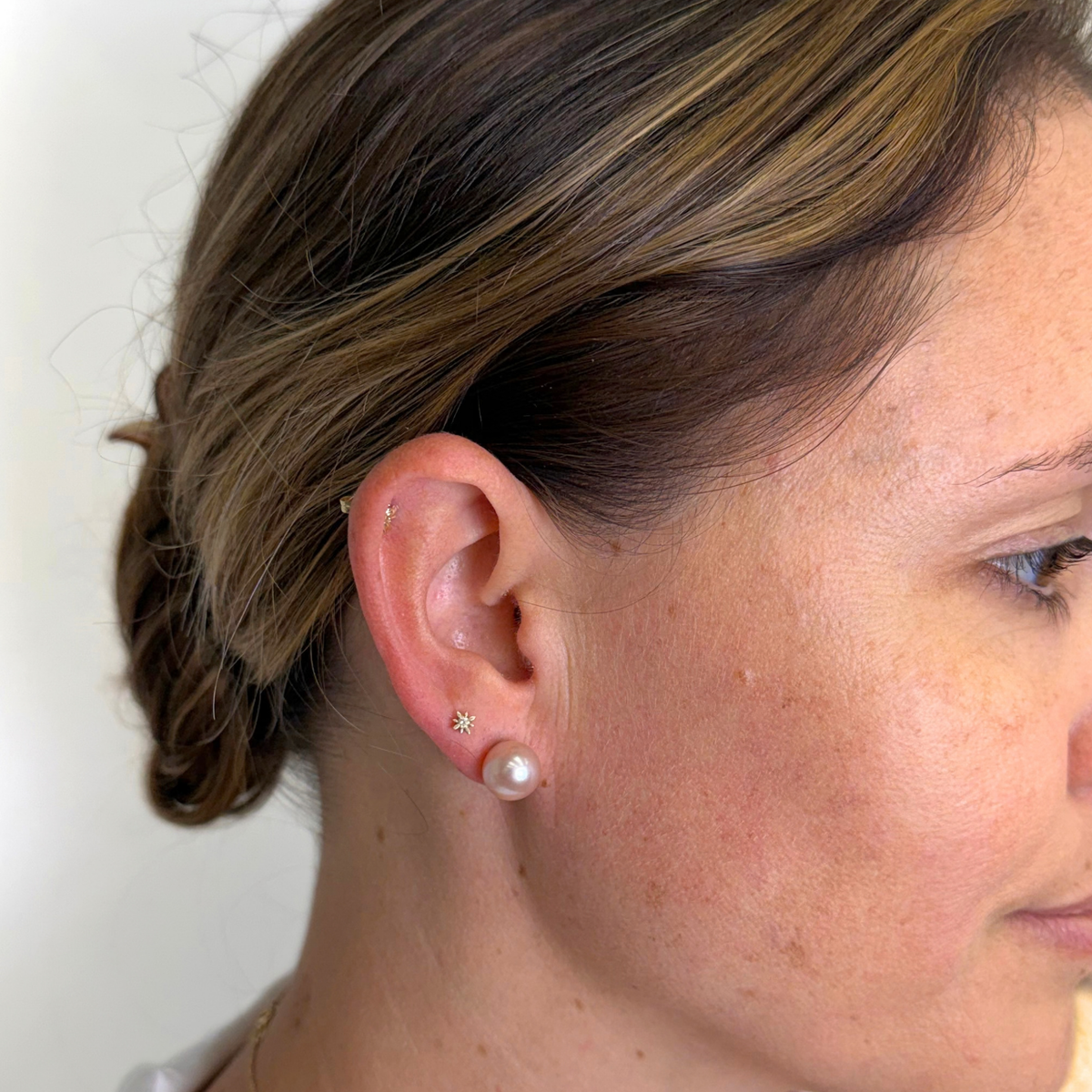 Close-up of a woman's ear wearing a small star-shaped stud earring and a pearl stud earring, with her hair pulled back and face partially visible in profile.