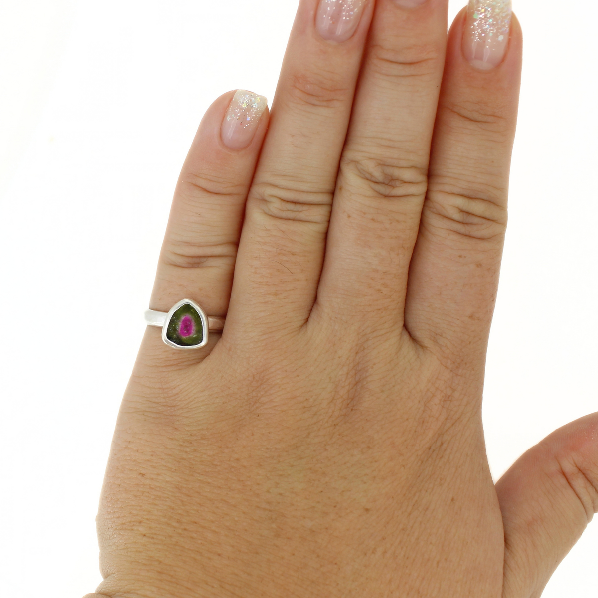 A silver ring with a triangular bezel holding a green stone with a pink center, shown worn on a person's ring finger against a white background.