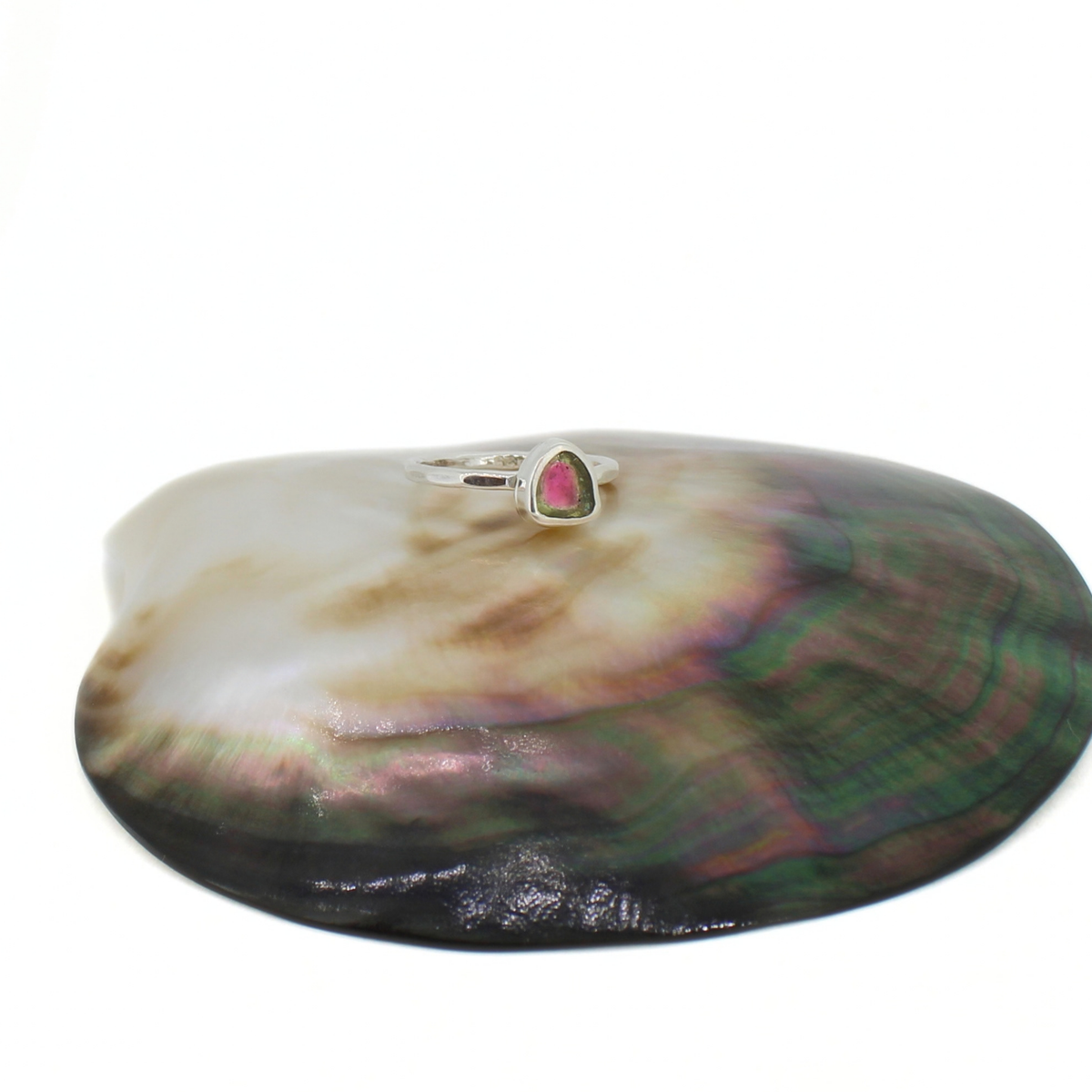 A silver ring with a small triangular-shaped pink and green watermelon tourmaline gemstone, displayed on top of a large iridescent abalone shell against a white background.