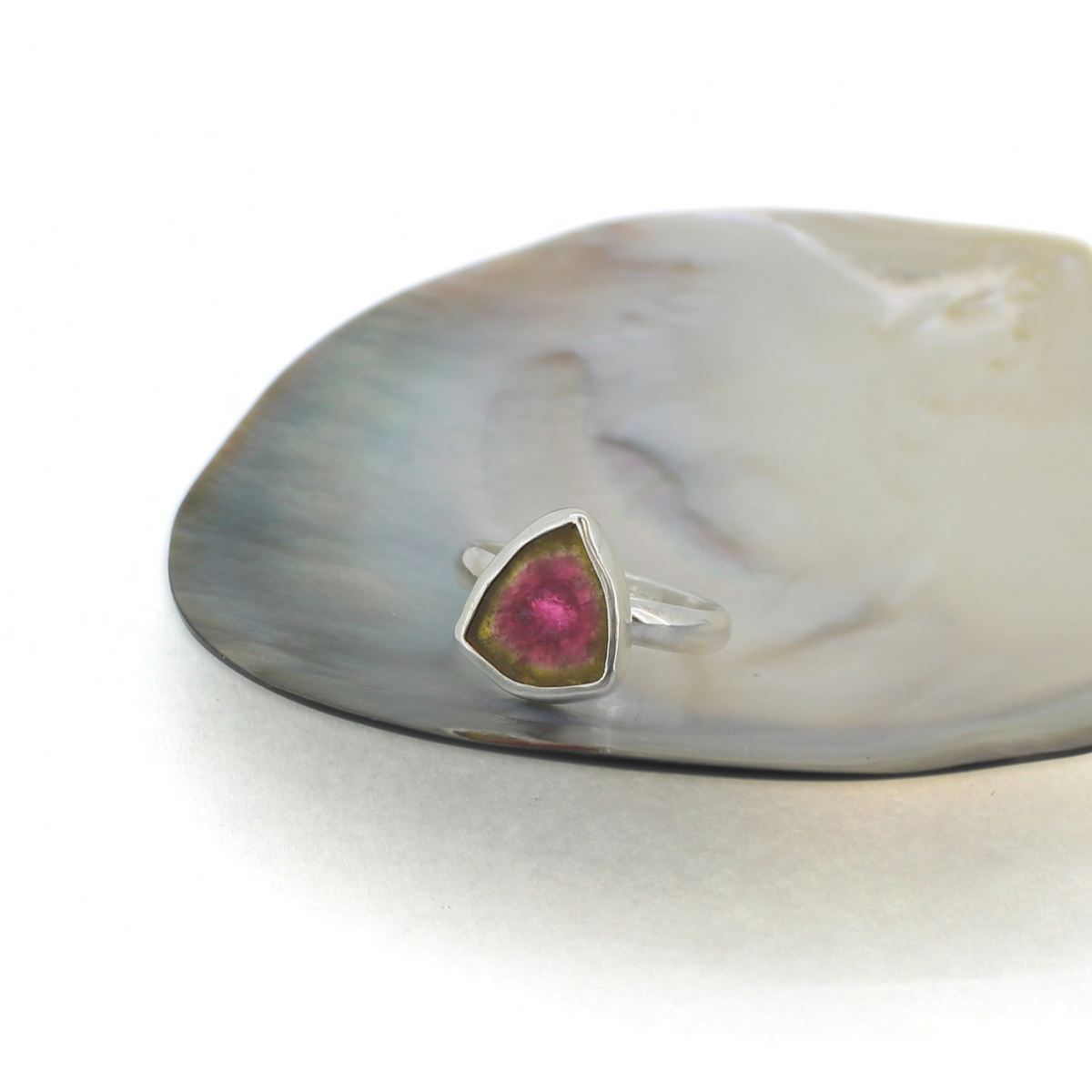 A silver ring featuring a triangular bezel-set pink tourmaline stone, displayed on a white and gray mother-of-pearl shell against a clean white background.