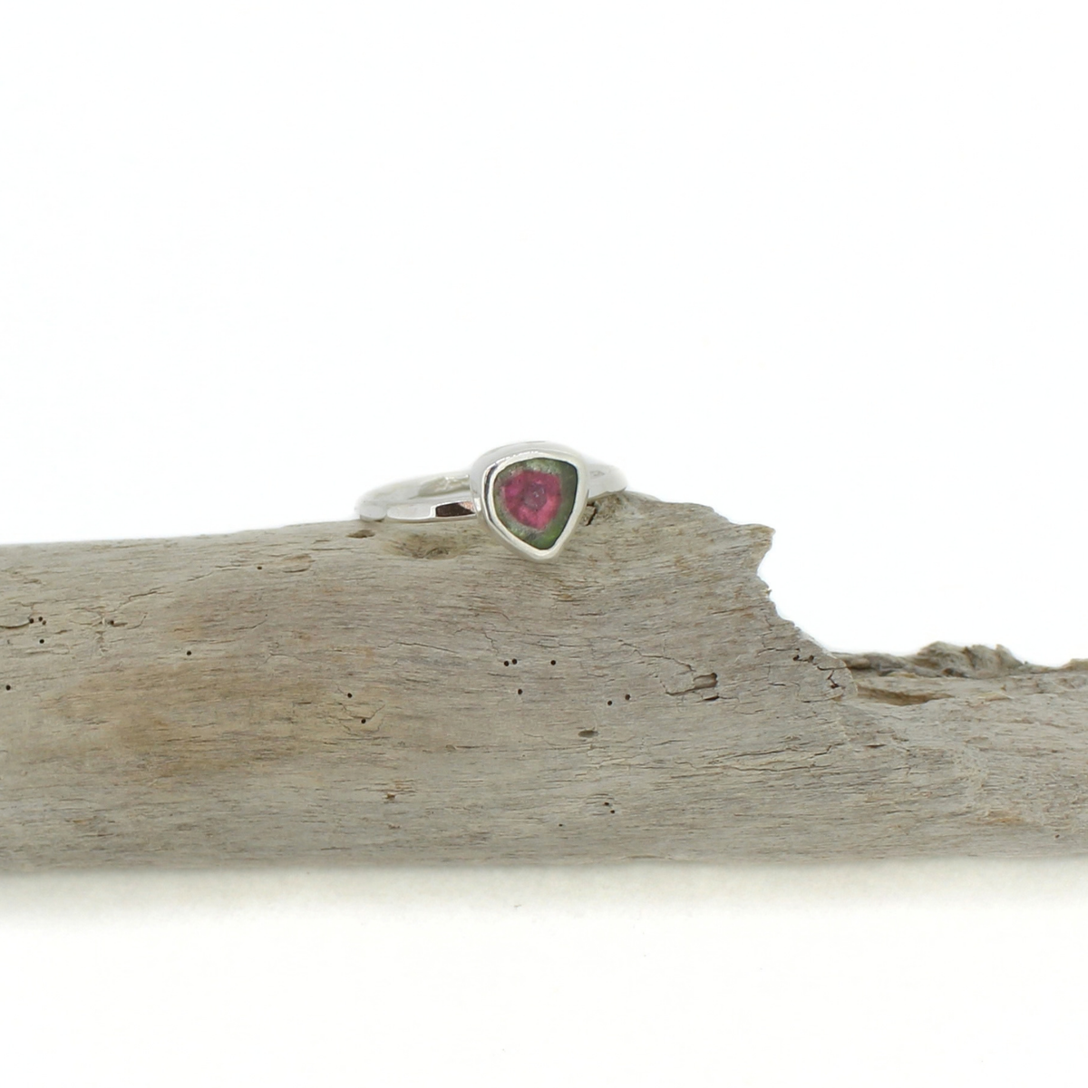 A silver ring with a small triangular-shaped pink and green watermelon tourmaline gemstone, displayed on a piece of weathered driftwood against a white background.